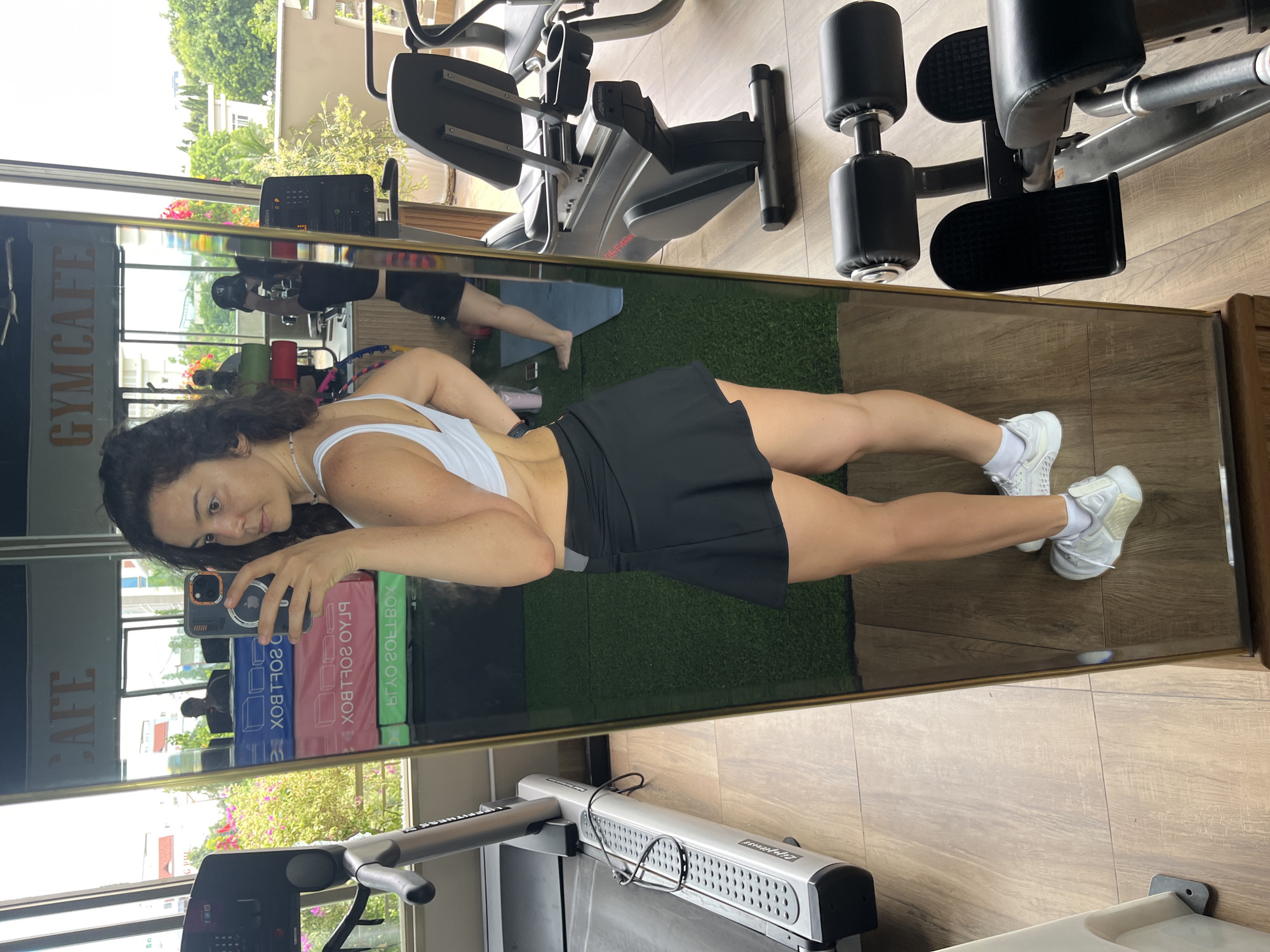 A woman is taking a mirror selfie in a gym cafe. She is wearing a white sports bra and a black skirt. She has dark curly hair and is looking at the camera. In the reflection of the mirror, we can see gym equipment, including treadmills and weight machines. There are also windows with a view of trees and buildings. The gym is decorated with signs that say "CAFE" and "GYMCAFE". The woman is standing on a wooden floor next to a treadmill. The overall scene is bright and airy, suggesting it is daytime. This gym cafe is located in the city of Phan Thiết, Vietnam.