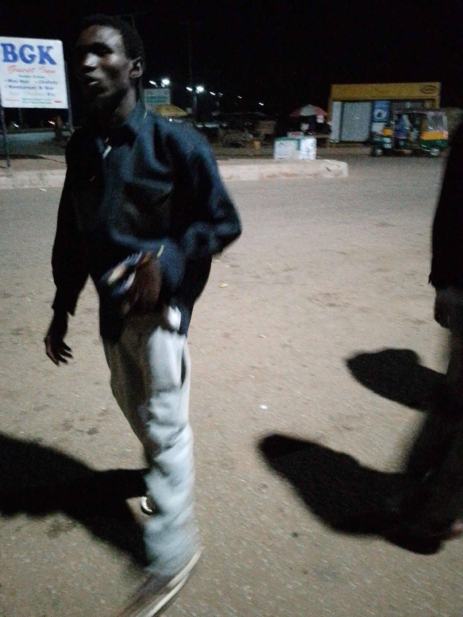 A dimly lit, outdoor scene at night. In the foreground, a young Black man wearing a dark collared shirt and light-colored pants walks towards the left of the frame. His body is slightly angled, and his left hand is raised near his chest. His face is visible and he appears to be speaking. The ground is paved and sandy. In the background, a white sign with blue text that reads "BGK Guest Inn" is visible on the left. Further back, there are buildings, streetlights, and three auto-rickshaws parked. The overall lighting suggests it is nighttime, and the shadows cast by the man and other objects indicate an artificial light source.