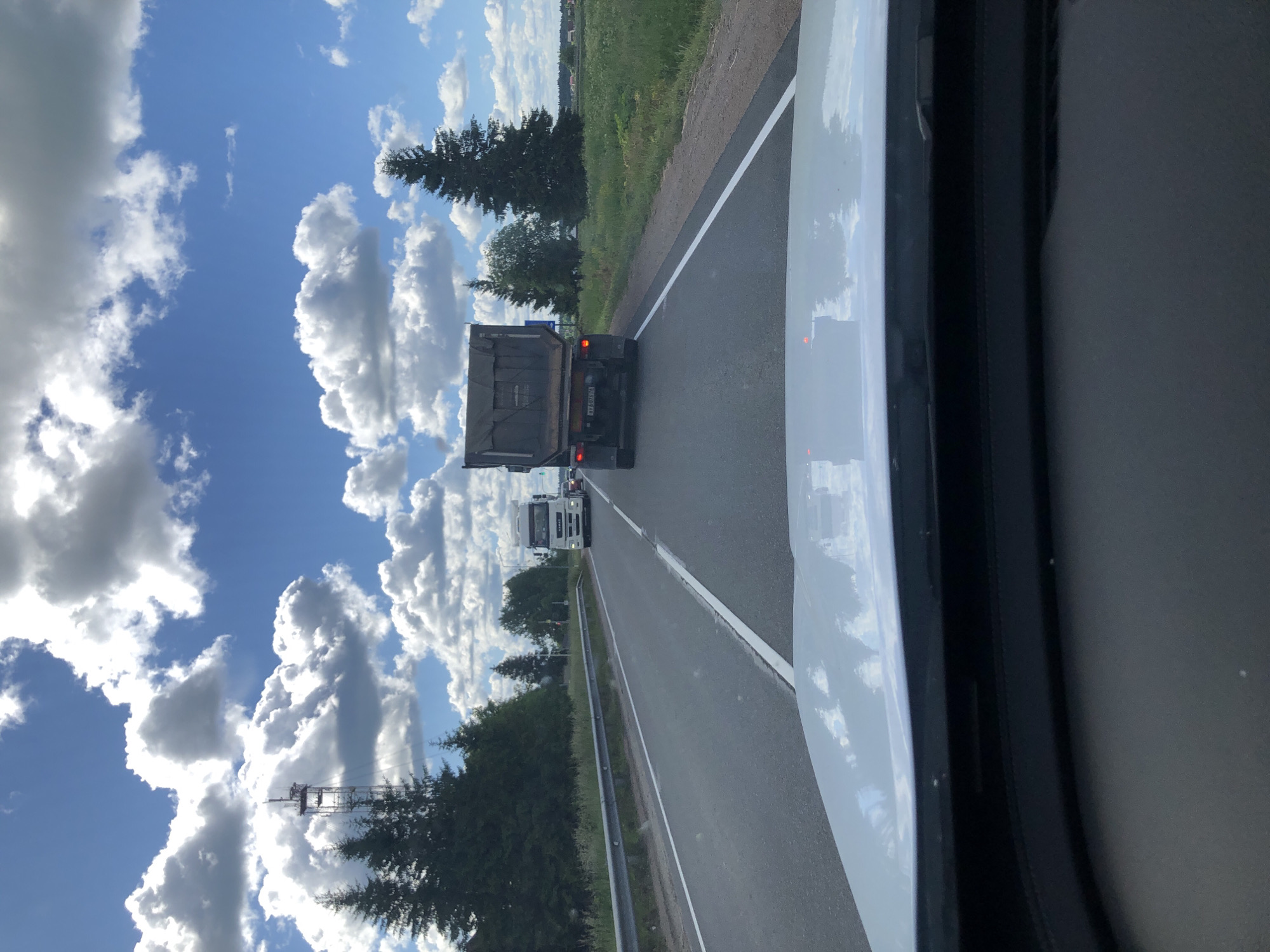 The image depicts a view from inside a vehicle traveling on a highway in the city of Dyatlitsy, Russia. The highway has two lanes in the direction of travel, separated by a white line. A guardrail runs along the left side of the road, and a grassy shoulder with trees borders the right. In the foreground, the white hood and windshield of the vehicle are visible.
Several trucks are present on the highway. The closest truck, in the right lane, is a dark-colored dump truck seen from the rear, with its license plate partially visible. Further ahead, in the left lane, a white semi-trailer truck is driving. The sky above is a bright blue, populated with fluffy white clouds.
The environment is rural, with dense green trees lining the road and fields of green grass and scattered houses visible in the distance on the right side. A tall communication tower with antenna elements is prominent in the left background, situated among the trees. The weather appears to be sunny with scattered clouds, suggesting a pleasant day.