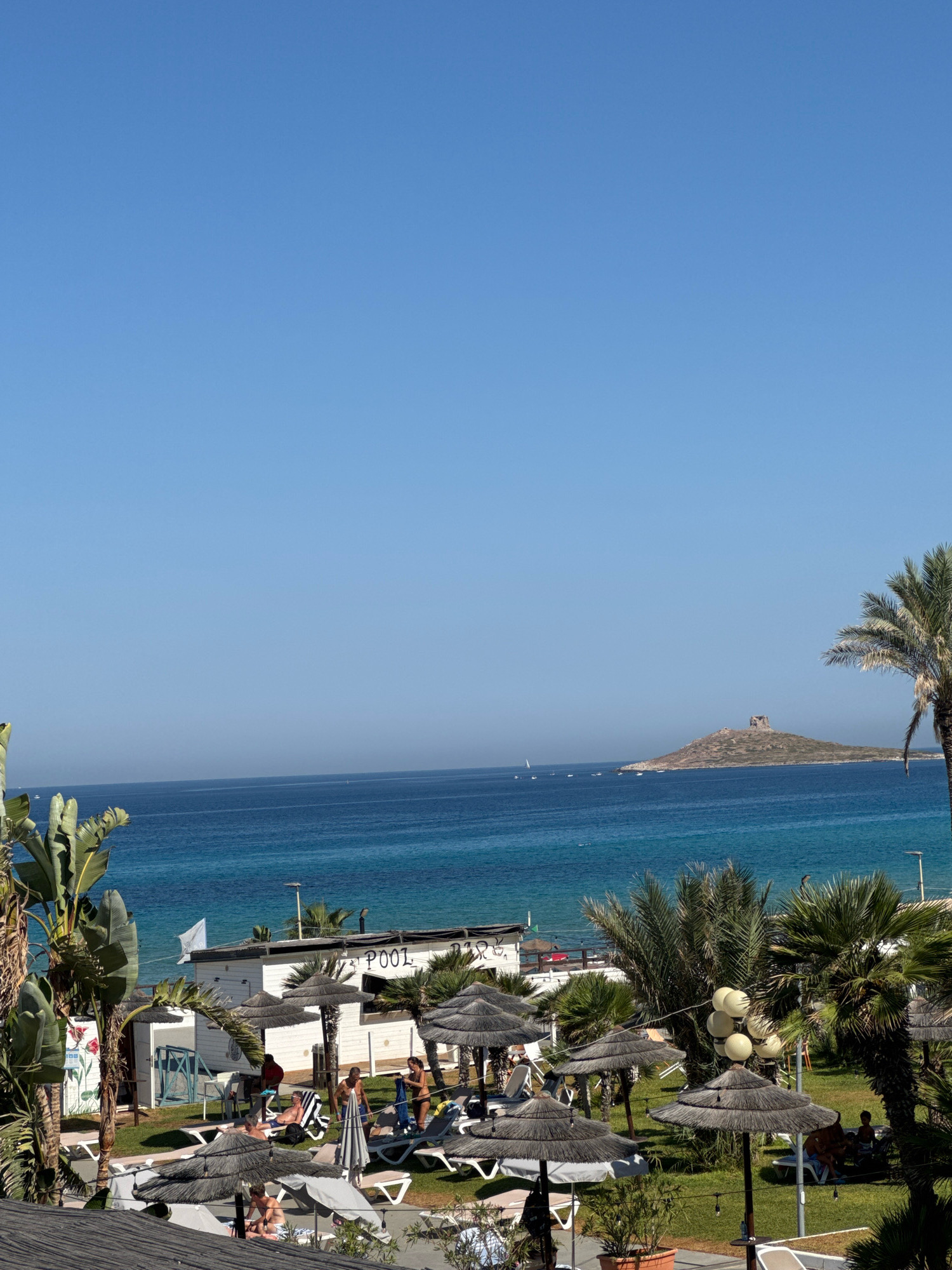 A high-angle shot captures a vibrant beach resort scene in Capaci, Italy, on a clear, sunny day. The deep blue sea stretches to the horizon, dotted with distant sailboats. In the middle ground, a white building with "POOL BAR" visible on its side stands amongst lush green grass and numerous thatched umbrellas shading lounge chairs. Several individuals are seen relaxing on the chairs, some interacting with each other. Palm trees and other tropical foliage are scattered throughout the resort area. Further out in the sea, a small island topped with ancient ruins is visible. The sky is a brilliant, cloudless blue, dominating the upper half of the frame.