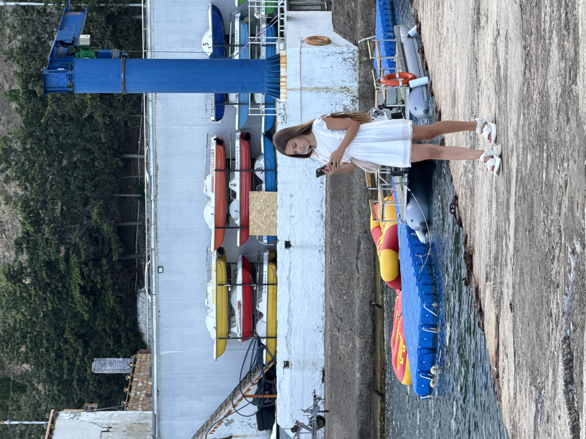 A young girl in a white dress and white sandals stands on a concrete pier, looking at her phone. She has long brown hair and a light-colored purse slung over her shoulder. In the background, a wall of white structures is lined with stacked paddle boats of various colors. To the right, a tall blue crane stands near the water. The water in front of the pier has several inflatable structures. The scene is set in Partenit, Ukraine, with a wooded hillside visible behind the wall.