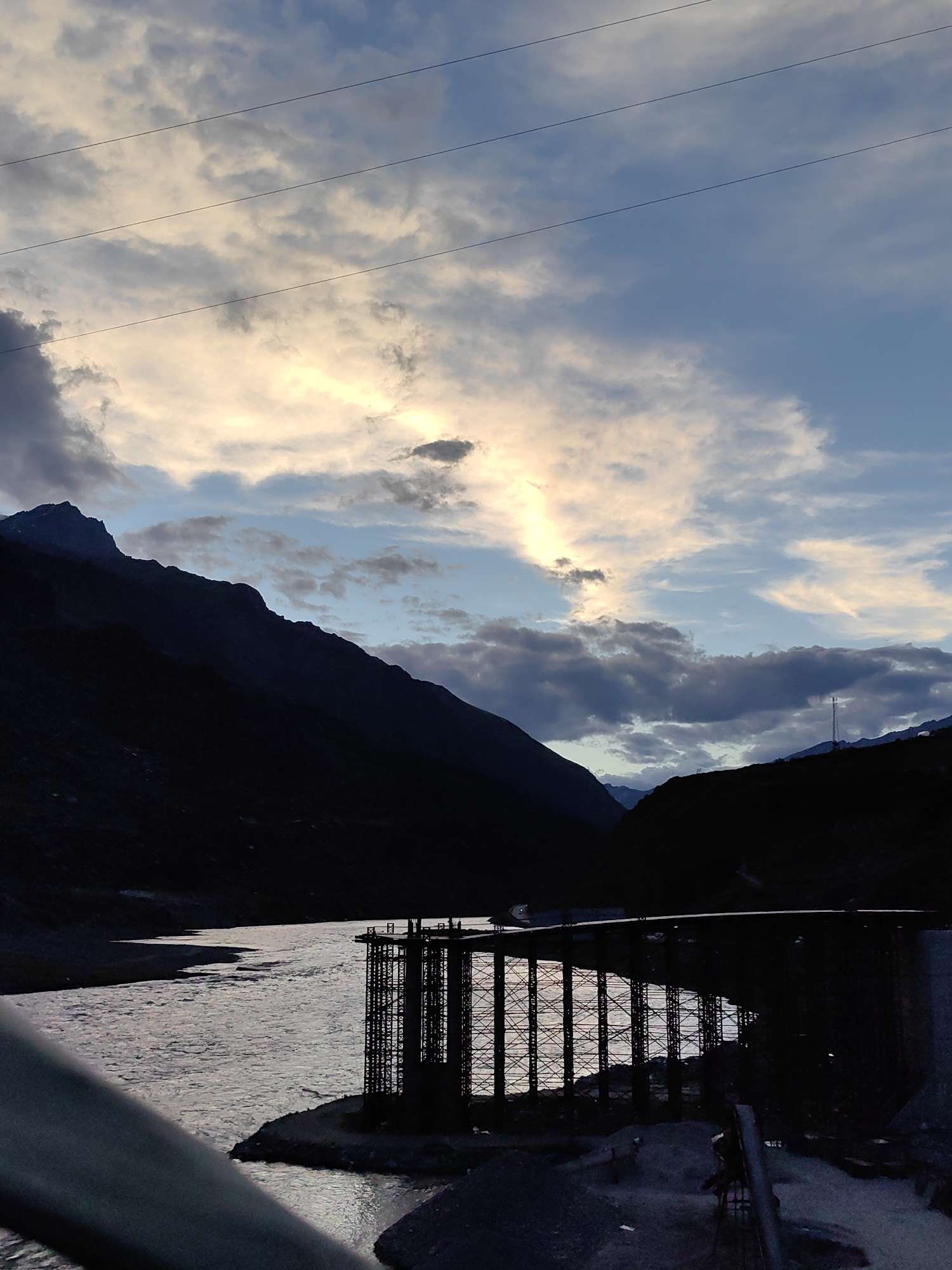 The image depicts a wide river flowing through a valley at dusk. On the right bank of the river, a bridge under construction is silhouetted against the water. The bridge's structure consists of numerous steel columns, creating a lattice-like appearance. The river's surface is illuminated by the reflection of the sky, which is filled with dramatic clouds. The left side of the image is dominated by the dark silhouette of a mountain range. Three parallel power lines stretch across the upper portion of the frame. The overall atmosphere is serene and majestic, with the fading light casting long shadows and highlighting the natural beauty of the landscape and the architectural elements of the bridge.
