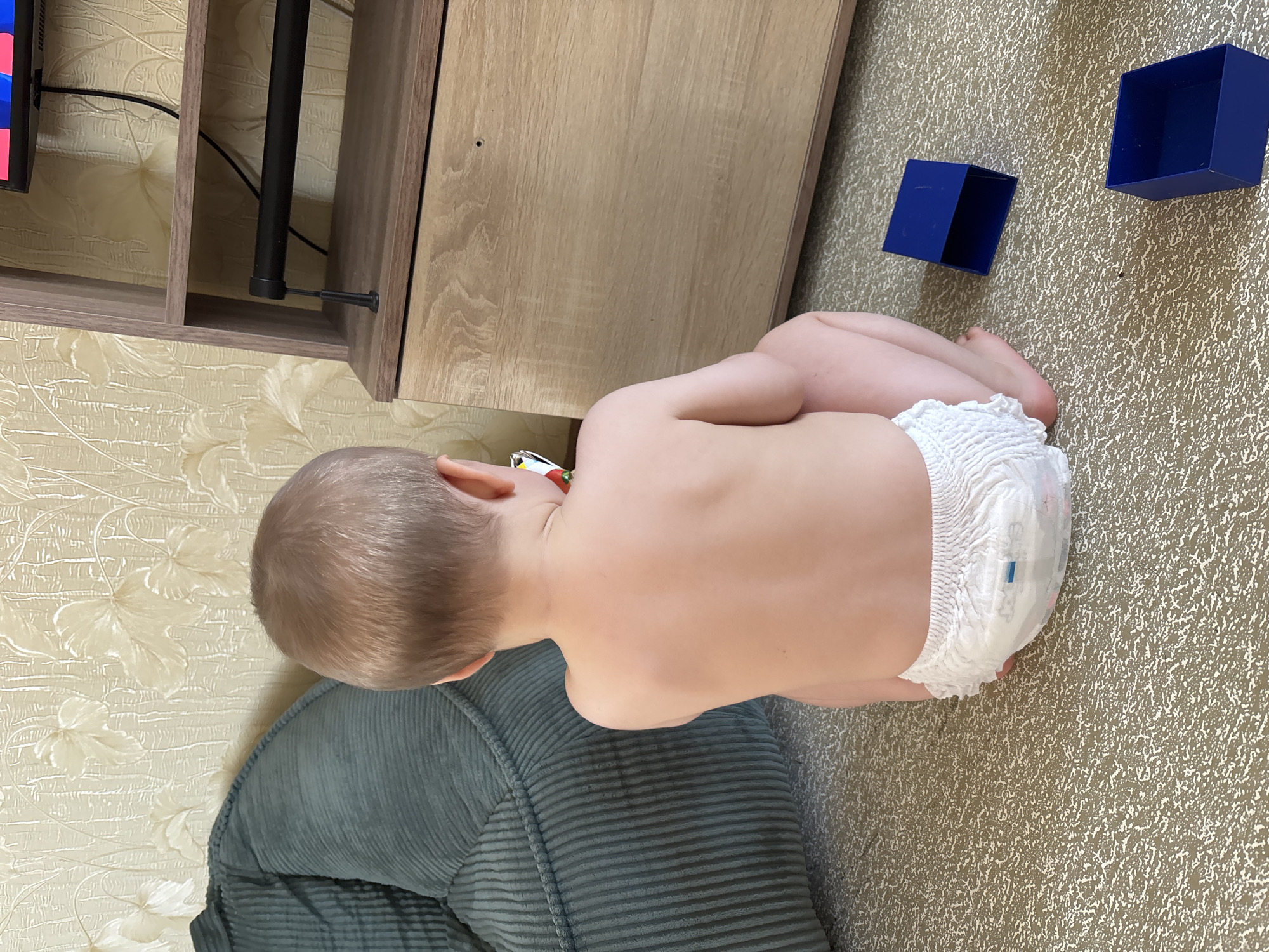 A toddler, viewed from the rear, sits on a carpeted floor in a room. The child wears a diaper and is facing away from the camera, towards a wooden media console with a television mounted above it. To the left of the toddler is a large, dark green beanbag chair. On the floor near the toddler are two open blue boxes. The wallpaper behind the child and furniture has a textured floral pattern in a light, neutral color. The lighting appears to be artificial, coming from above and slightly to the right, casting soft shadows. This scene is situated within a residential dwelling in Naberezhnye Chelny, Russia.