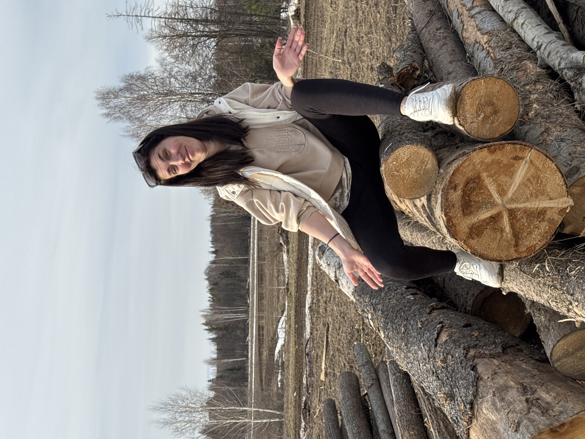 A woman with dark hair wearing black leggings, white sneakers, and a light-colored hoodie and jacket is sitting on a pile of logs. The setting appears to be a field in Van'ki, Russia, with trees and some remaining snow visible in the background. The woman is gesturing with her hands while balancing on the logs.