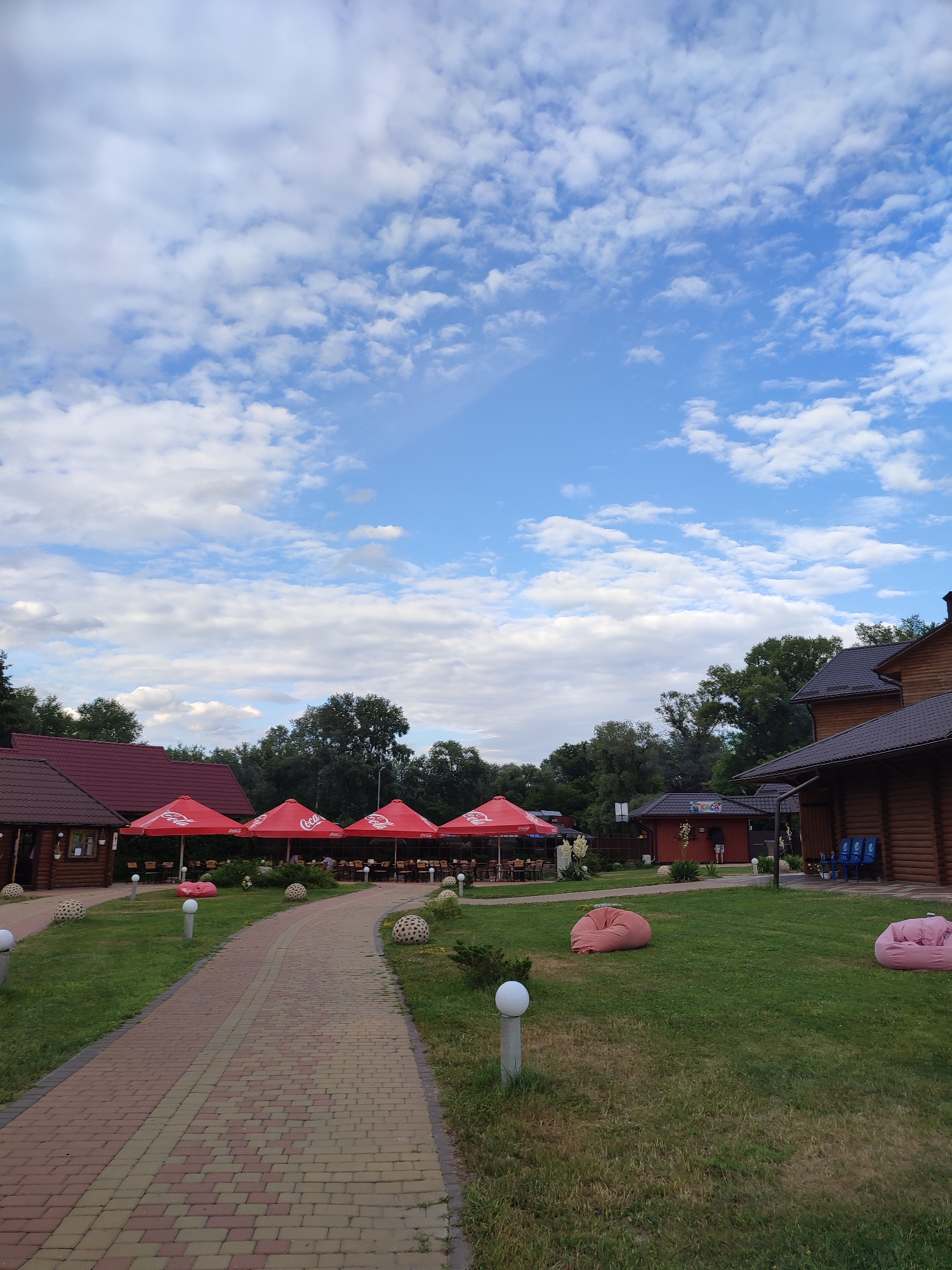 The image depicts an outdoor seating area of a restaurant or resort. A paved walkway curves through a grassy area, leading towards several wooden buildings with red roofs. Lined up in front of one of the buildings are numerous red umbrellas, under which tables and chairs are set up for dining. The umbrellas bear the "Coca-Cola" logo.
In the grassy area, there are decorative spherical objects and modern lampposts. Two large pink beanbag chairs are visible on the lawn, suggesting a relaxed atmosphere. The background is filled with lush green trees and foliage.
The sky is a vibrant blue with scattered white clouds, indicating pleasant weather, likely daytime. The architecture of the buildings, with their wooden log construction and dark roofs, suggests a rustic or traditional style. There is a small building with "KACA" written on it.
No specific activity or event is clearly depicted, but the setup implies a dining or leisure setting. The overall mood is tranquil and inviting. The location is identified as Shcherbani, Ukraine.