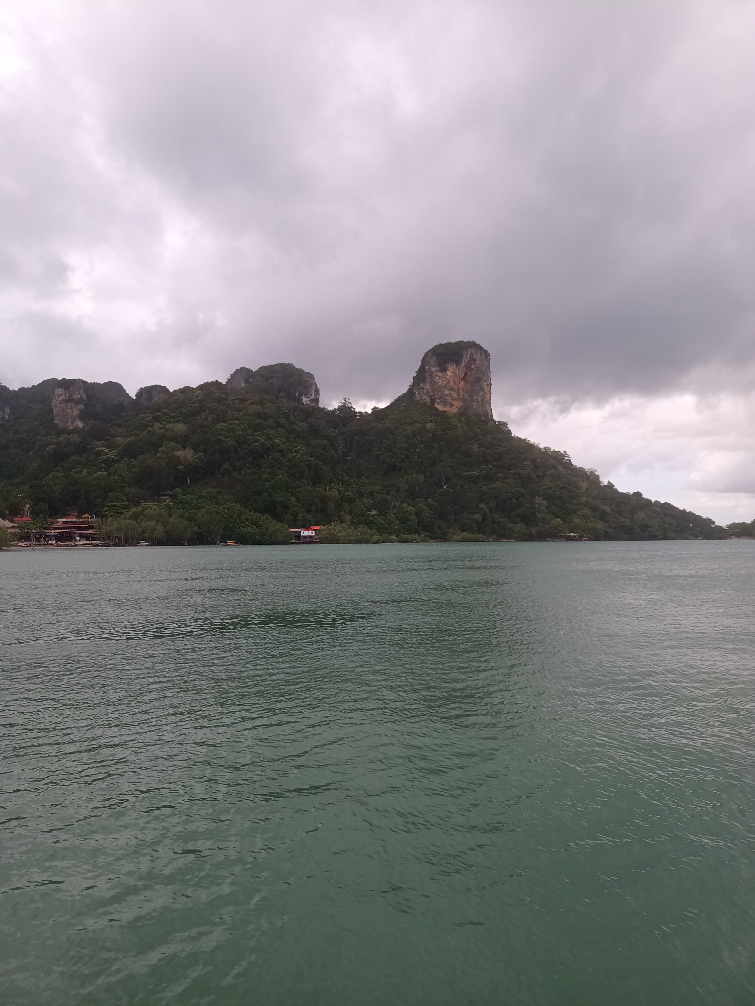 The image depicts a seascape in Ban Ao Nang, Thailand, under an overcast sky. The foreground is dominated by green-tinted water with gentle ripples. In the midground, a landmass is covered with dense green vegetation and rocky cliffs. Buildings are seen along the coast. A two story structure with red roof and white walls is sitting in the water near the shoreline. The background is a heavy, grey sky.