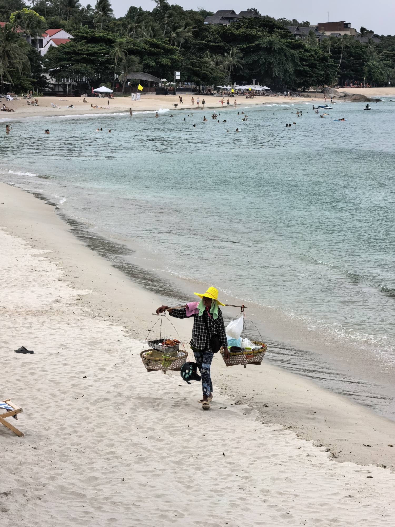 A person walks along a sandy beach carrying a yoke across their shoulders, from which hang two baskets. The basket on the right appears to contain food items, possibly grilled. The basket on the left holds various items, including what looks like produce and plastic bags. The person wears a plaid shirt, patterned pants, and a bright yellow sun hat. They are walking on the wet sand near the water's edge.
The beach is populated with other people enjoying the ocean. Some are swimming or wading in the clear turquoise water, while others are sunbathing on lounge chairs or walking along the shore. In the background, a lush green hillside is covered with trees and buildings, suggesting a tropical resort area. A few small structures, possibly beach bars or restaurants, are visible along the coastline, with umbrellas providing shade. The water is calm with gentle waves lapping the shore. The sky appears overcast, indicating a cloudy day, but the light is diffused, suggesting it might be daytime, possibly late morning or afternoon. The overall scene conveys a sense of relaxation and typical beach activity. There is no visible text in the image.