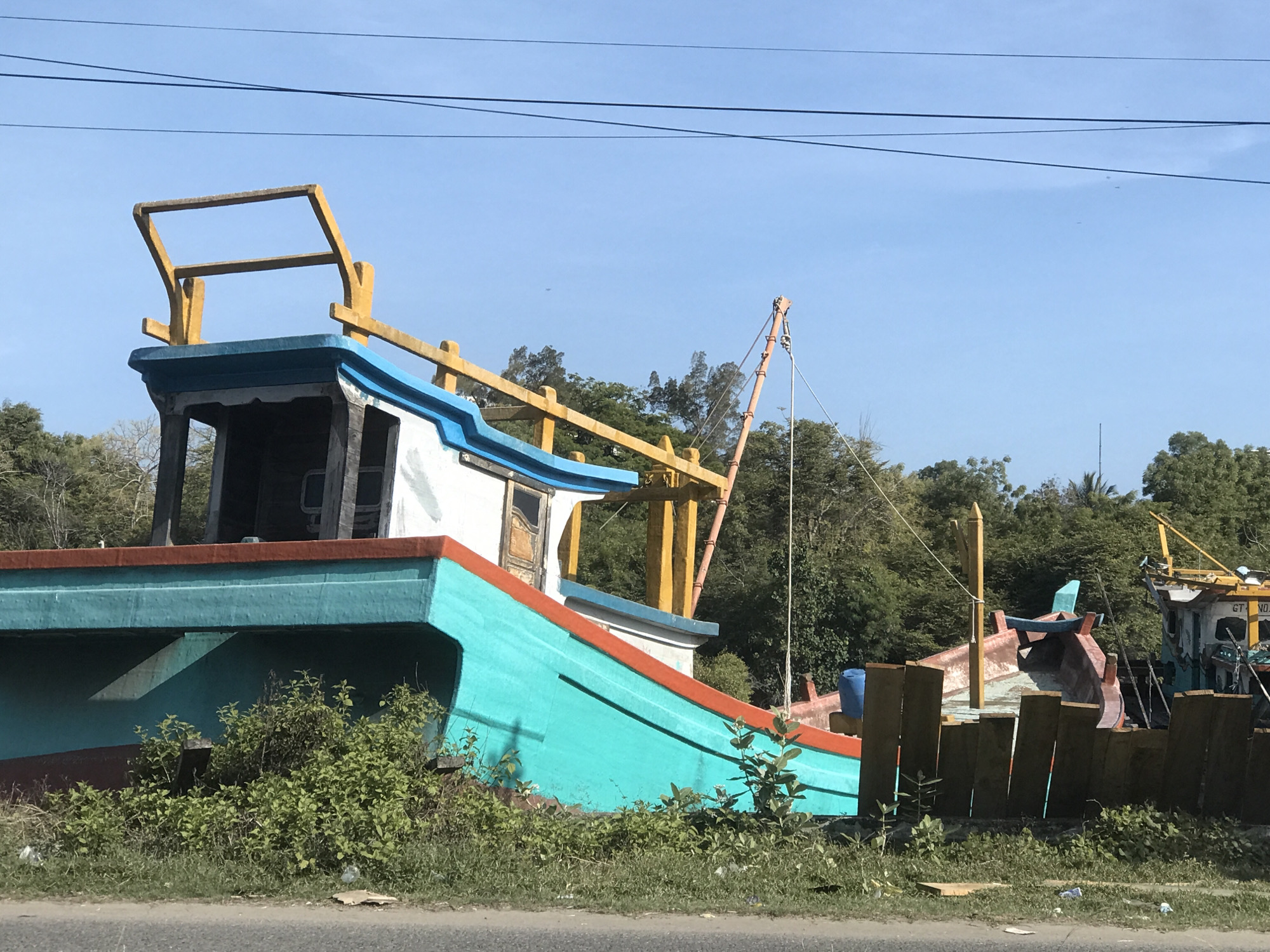 The image shows an abandoned or decommissioned colorful fishing boat in what appears to be a yard or storage area. The boat is painted in turquoise, white, red, and blue with yellow structural components. Vegetation, including grasses and bushes, grows around the base of the boat. Other boats are also partially visible in the background and wood planks. The sky is blue with overhead power lines. The location is implied to be Lhokseumawe, Indonesia, but there is no location tag or any writing on the boats.