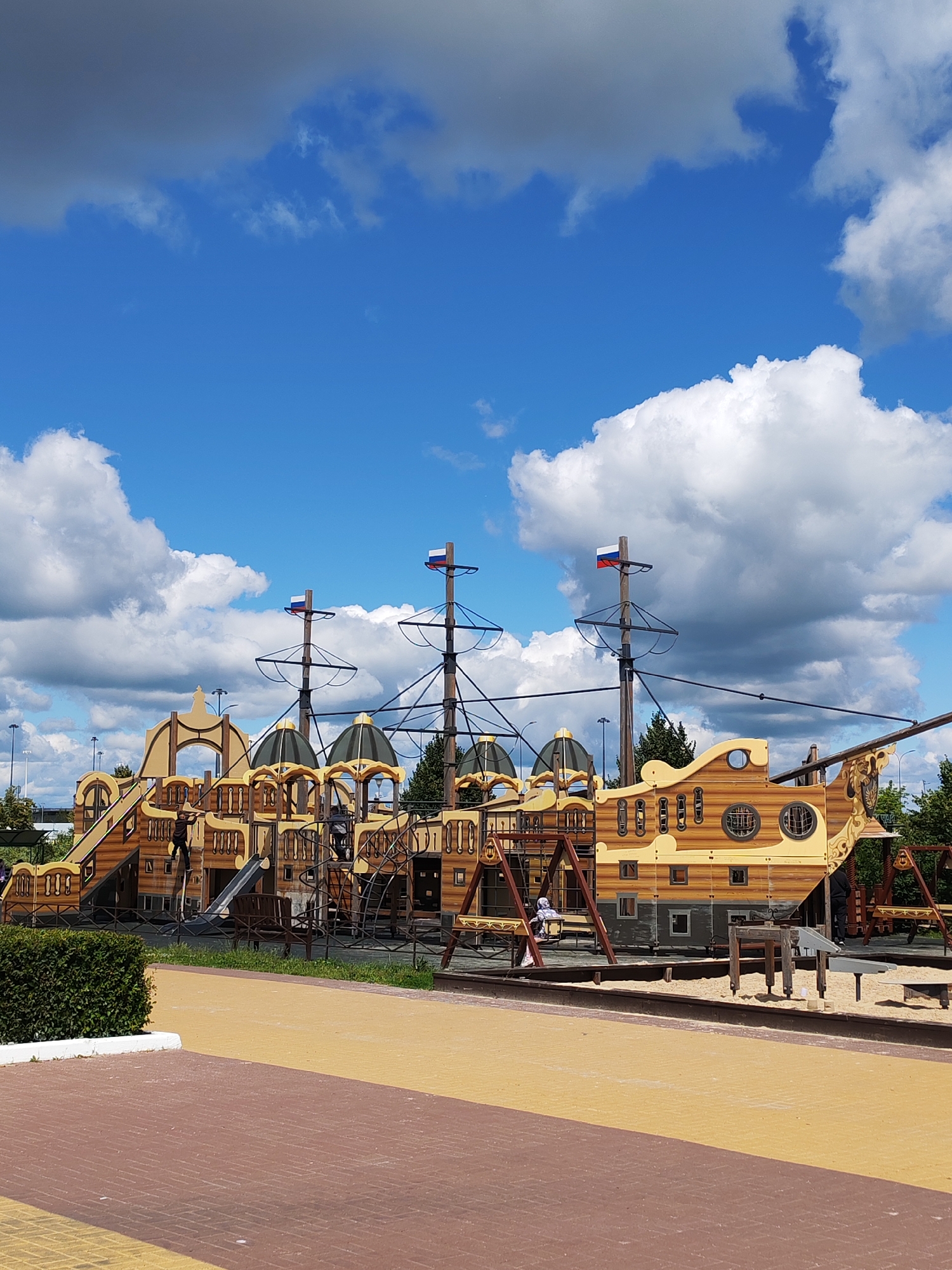 The image shows a large, wooden pirate ship-themed playground on a sunny day with scattered white clouds against a bright blue sky. The playground is elaborate, featuring multiple levels, slides, climbing structures, and what appear to be swings. Russian flags are visible on the masts of the ship.
In the foreground, there are pathways made of red brick and yellow paving stones. To the left, a neatly trimmed green hedge is visible. The playground itself is the central focus, with its intricate design that mimics a galleon. The ship's structure is made of light brown wood with darker brown detailing and various windows and openings.
Several people are present. One person, wearing dark clothing, is climbing on the structure. Another person, dressed in light-colored clothing, is sitting on a swing. In the distance, other figures are visible on the playground, suggesting it's in use. The scene appears to be a public park or recreational area, designed for children's play.
The time of day is daytime, likely late morning or early afternoon, given the bright sunlight casting shadows. The weather is pleasant, with a clear blue sky and fluffy clouds, indicating fair weather. The presence of Russian flags suggests the location is in Russia, and the prompt specifies Nizhny Novgorod. There is no visible text in the image.