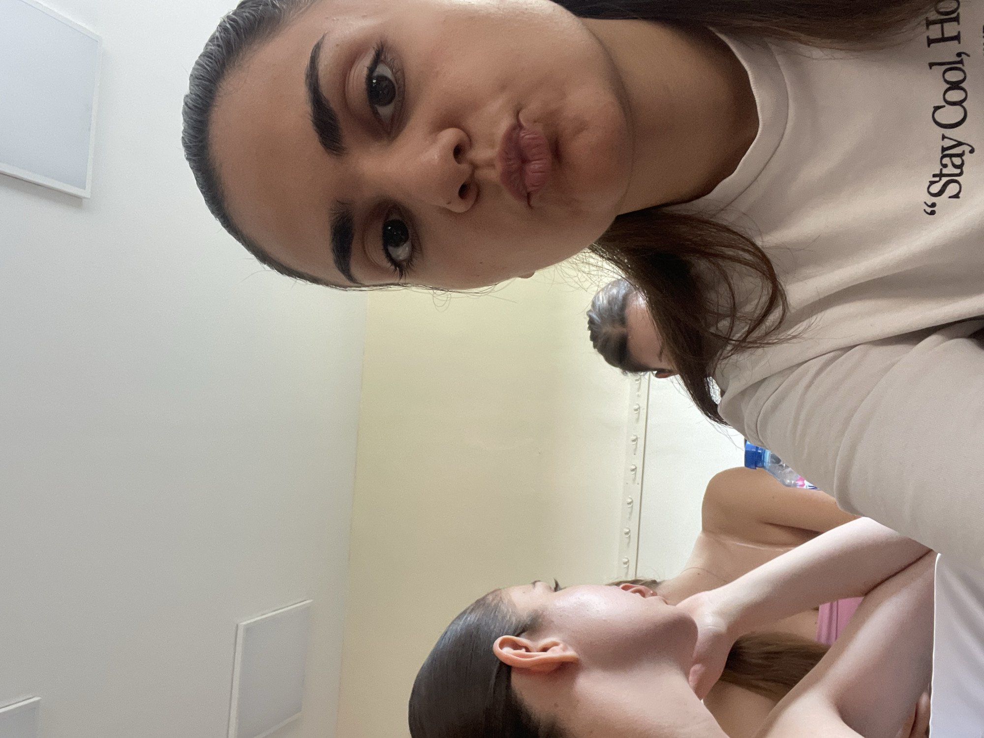 The image shows a selfie of a woman indoors. She is wearing a white t-shirt with the text "Stay Cool, Home." The background appears to be a room with white walls. In the reflection, another person is visible with arms on the other person's neck. There is no indication of location within Burgas, Bulgaria.
