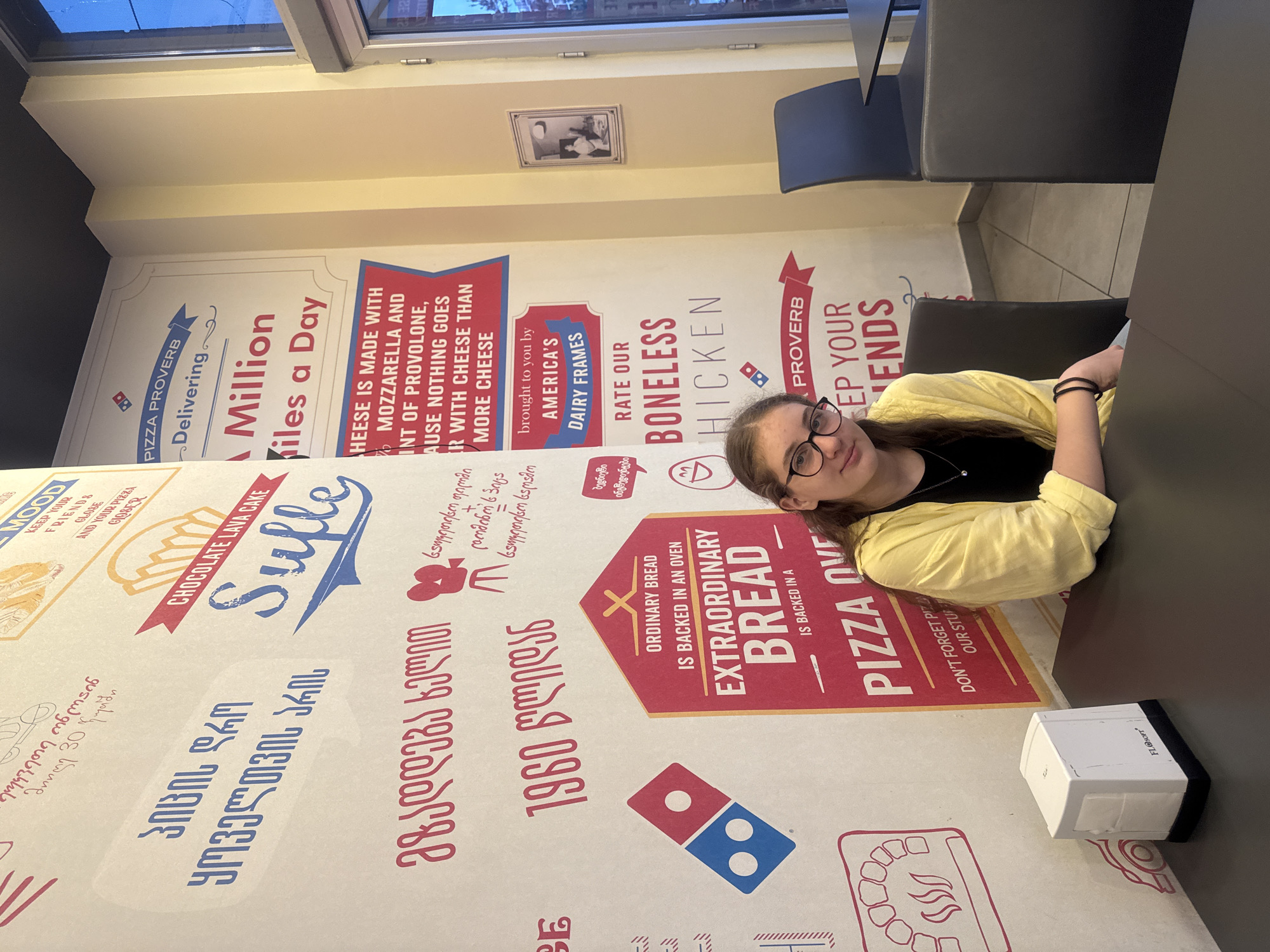 In a brightly lit, casual dining establishment in Tbilisi, Georgia, a young woman with dark hair, wearing glasses and a yellow cardigan over a black top, is seated at a table. The walls of the establishment are adorned with a vibrant mural featuring text in Georgian and English, alongside Domino's Pizza branding. Prominently displayed are slogans like "PIZZA PROVERB: Delivering a Million Miles a Day" and "CHEESE IS MADE WITH MOZZARELLA AND POINT OF PROVOLONE, BECAUSE NOTHING GOES BETTER WITH CHEESE THAN MORE CHEESE." Another section highlights "ORDINARY BREAD IS BACKED IN AN OVEN FOR EXTRAORDINARY BREAD." On the table in front of the woman is a white napkin dispenser. The atmosphere suggests a modern, fast-casual restaurant, possibly a Domino's Pizza branch.