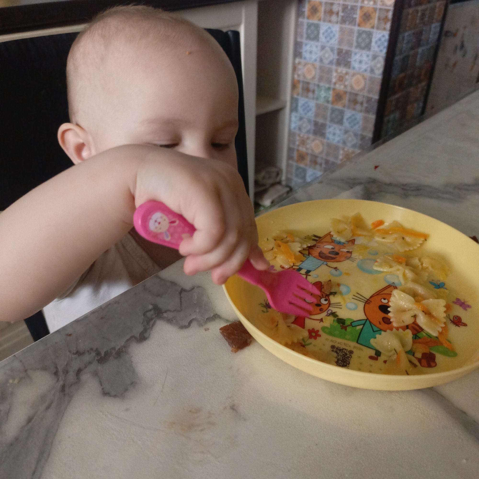 The image provides a close-up view of a baby or toddler engaged in eating. The child's right arm and hand dominate the center-left, firmly grasping a bright pink child-sized fork. A cartoon rabbit sticker adorns the handle of the fork. The child's head, with fair skin and light hair, is partially visible on the left, looking downwards towards the food.
A round, yellow child's plate, decorated with cartoon cat characters and butterflies, sits on a marbled table surface. The plate contains bowtie pasta (farfalle) mixed with small orange pieces of what appear to be carrots, and a light sauce. Some pasta is on the fork, indicating the child is actively trying to eat. To the left of the plate, on the table, is a small, reddish-brown rectangular food item, possibly a fruit chew or a piece of dessert. The table itself has a grey and white marbled pattern with some darker veins and faint streaks of brown/yellow.
In the background, a dark chair or highchair back is visible behind the child. Beyond that, there's a white shelving unit, and a wall covered in decorative tiles featuring a checkered pattern of blue, orange, brown, and white squares. The setting appears to be an indoor kitchen or dining area.
The primary activity is the baby attempting to self-feed. The child's face is mostly obscured, but the downward gaze suggests concentration on the task of eating. The time of day and weather are not discernible, as the scene is indoors with artificial or ambient light. On the yellow plate, near the bottom left cat, a small, dark, pixelated graphic, possibly a QR code, is visible, along with very faint, tiny text that includes fragments like "AD CTC" and "2024".