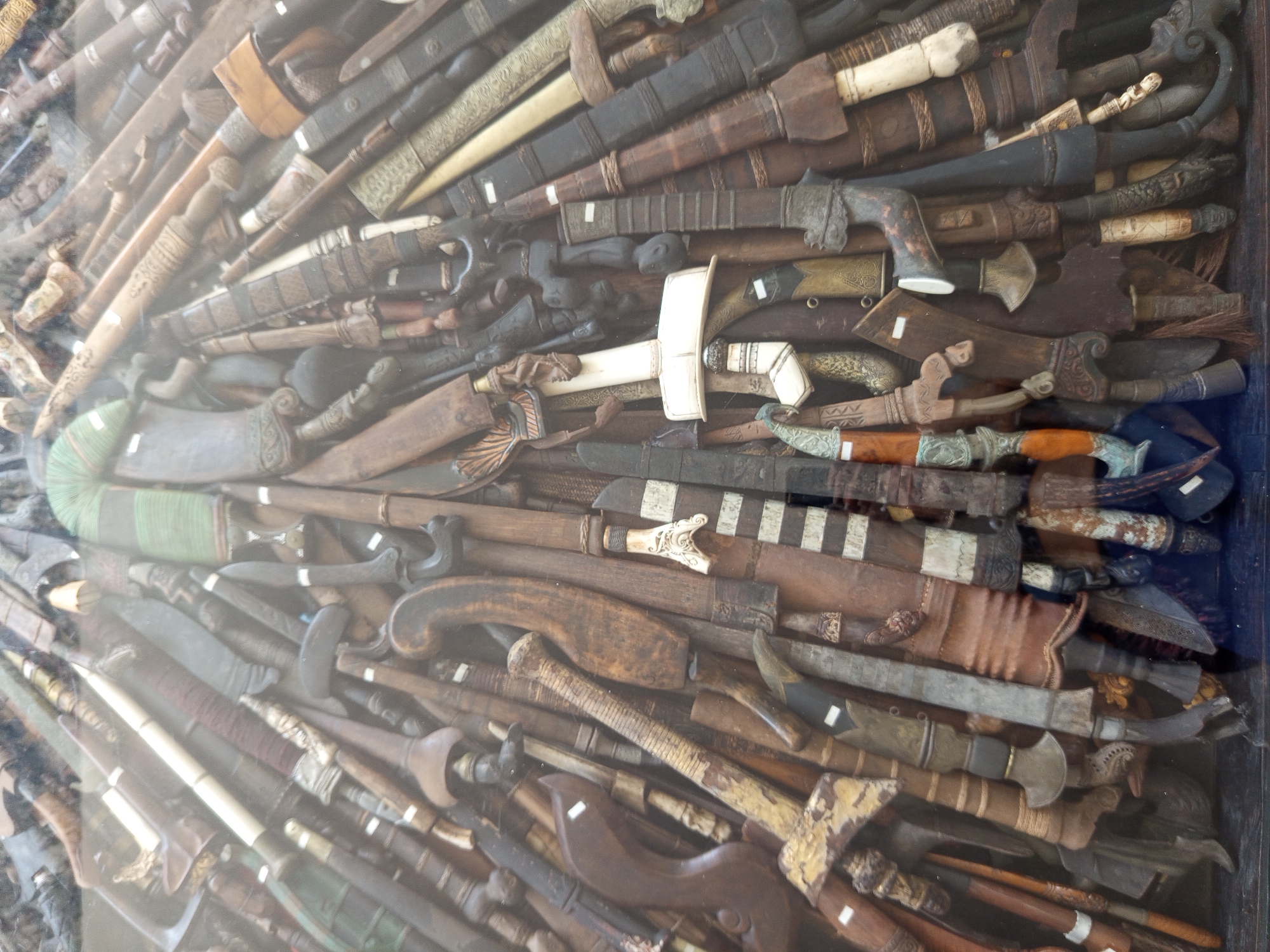 The image shows a collection of Indonesian edged weapons, primarily swords and daggers, displayed behind glass. The blades and scabbards vary in size, shape, and material, exhibiting diverse wood tones, ivory, and metalwork. Many handles are intricately carved. The scene is likely a museum display or a private collection. North Kuta, Indonesia, is given as the location.