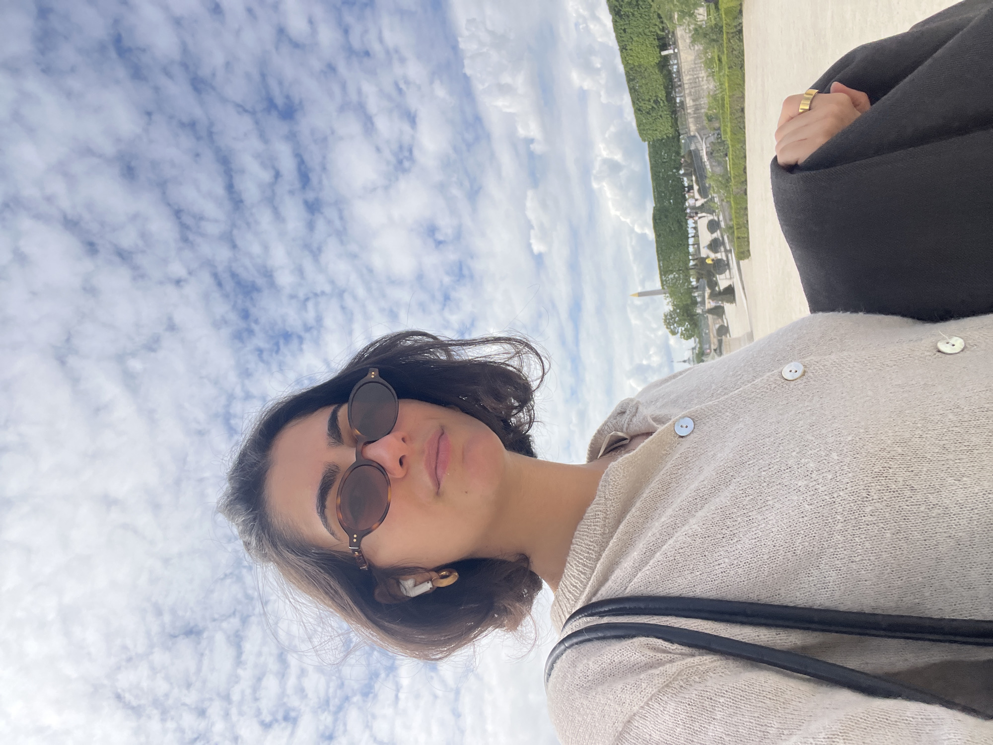 A selfie taken from a low angle captures a person looking upwards. The individual, appearing to be female, has dark, short hair and wears round, tortoiseshell-patterned sunglasses. A white earbud is visible in their right ear, accompanied by a small gold-toned hoop earring. They are dressed in a light-colored, possibly beige, knitted cardigan with small, round, white buttons. A dark strap, likely from a bag, crosses their torso. A hand, adorned with a wide gold ring on the index finger, holds a piece of dark fabric, possibly a jacket or garment.
The background is largely composed of a bright blue sky with scattered white, cumulus clouds. To the right of the subject, a formal garden with neatly trimmed green hedges and trees is visible, extending into the distance. A notable landmark in the background is a tall, slender obelisk, which strongly suggests the Obelisk of Luxor in Place de la Concorde, Paris. The bright daylight indicates the photo was taken during the day in clear weather, likely in or near the Tuileries Garden, with the Parisian landmark providing a distinctive location cue. No discernible text is visible.