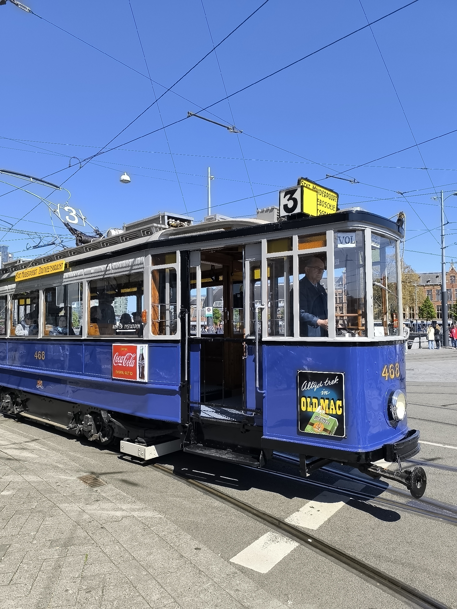 The image shows a blue tram in Amsterdam, Netherlands. The tram is marked with the number 468 and route number 3. The tram has advertisements for "Coca-Cola" and "OLD MAC" cigarettes. The route number sign on top of the tram indicates it travels to "STAT MUIDERDOORT", "EBOSCHJE", and "SGD". The route number sign on the side indicates the tram travels to "STAT MUNDEDDOORT ZOUTNEETSGRACHT". A driver wearing a suit is operating the tram. Other people are visible inside the tram and on the sidewalk near the tram. Overhead electrical wires for the tram are visible against a clear blue sky. The ground is paved with stone blocks.