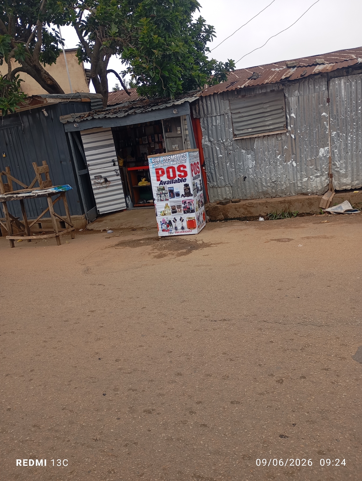 In an outdoor scene at an unknown urban location, a small storefront is visible. The building is constructed with corrugated metal and has a tin roof. In front of the store, a display box showcases various cosmetic and herbal medicine products. A sign on the box reads "SUFI COSMETICS AND HERBAL MEDICINE POS Available" with a phone number "TEL: 09035842057". A wooden table and chair set are placed to the left of the storefront. The ground is paved with asphalt. Power lines are visible overhead.