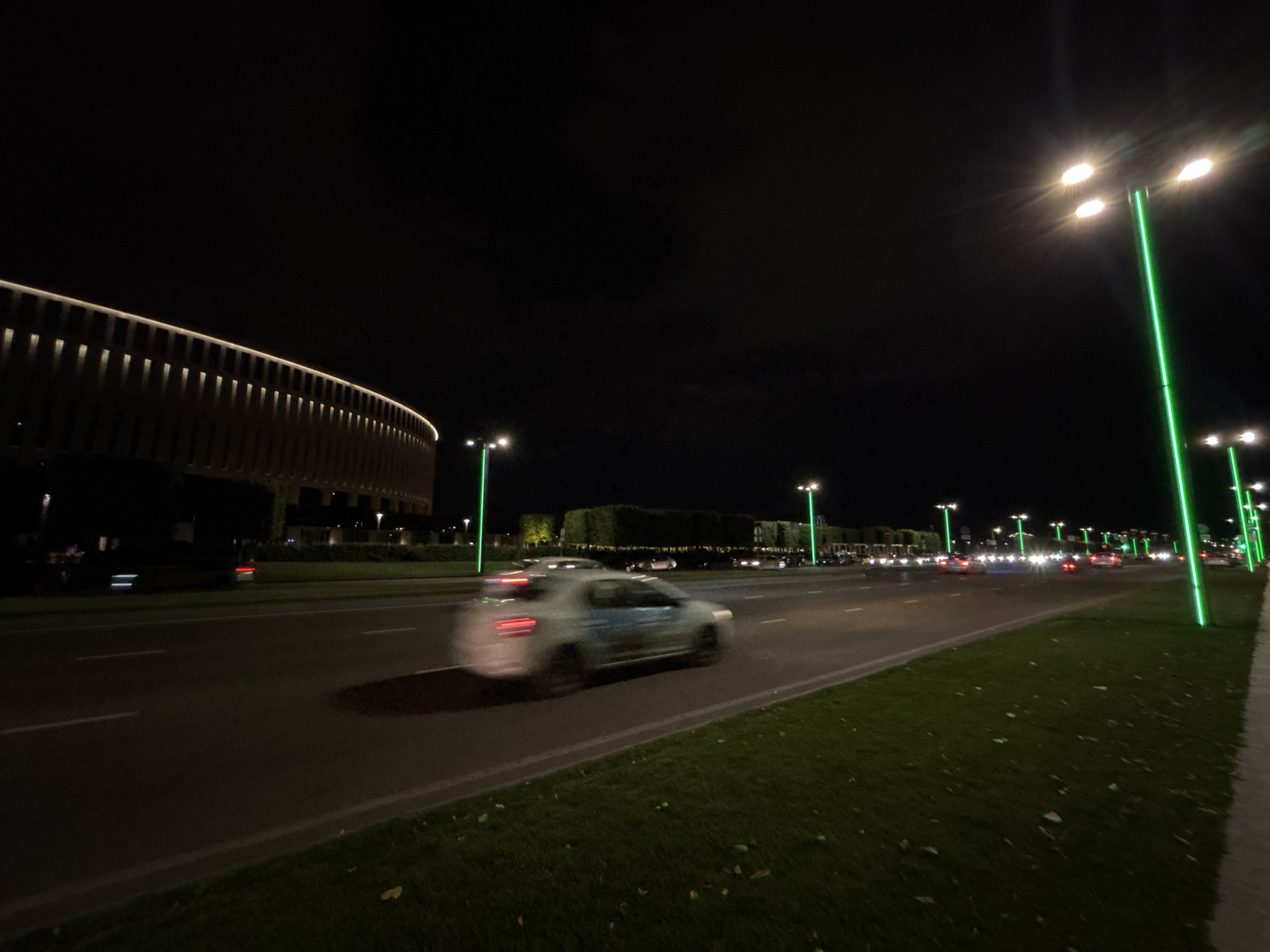 This is a nighttime photograph of a wide, multi-lane road in Krasnodar, Russia. The scene is illuminated by streetlights, some of which are a distinctive bright green. On the left side of the road, a large, curved building with a modern design is visible, its facade lit with a row of warm lights. The building appears to be a stadium or arena, characterized by its arched structure and a ribbed or panelled exterior.
The road is wet, suggesting recent rain or a damp evening. Several cars are driving on the road, some captured with motion blur due to the long exposure of the photograph. One car in the foreground is particularly blurred, showing streaks of light from its headlights and taillights. Further down the road, more cars are visible, their headlights creating bright points of light.
The right side of the road features a well-maintained grassy area with more green streetlights lining the sidewalk or pathway. The grass is dark, with some scattered fallen leaves. The sky is dark and overcast, typical of nighttime. There is no visible text in the image. The overall atmosphere is one of urban nighttime activity, with the modern architecture and bright streetlights creating a slightly futuristic feel. The time of day is clearly night, and the weather appears to be damp.