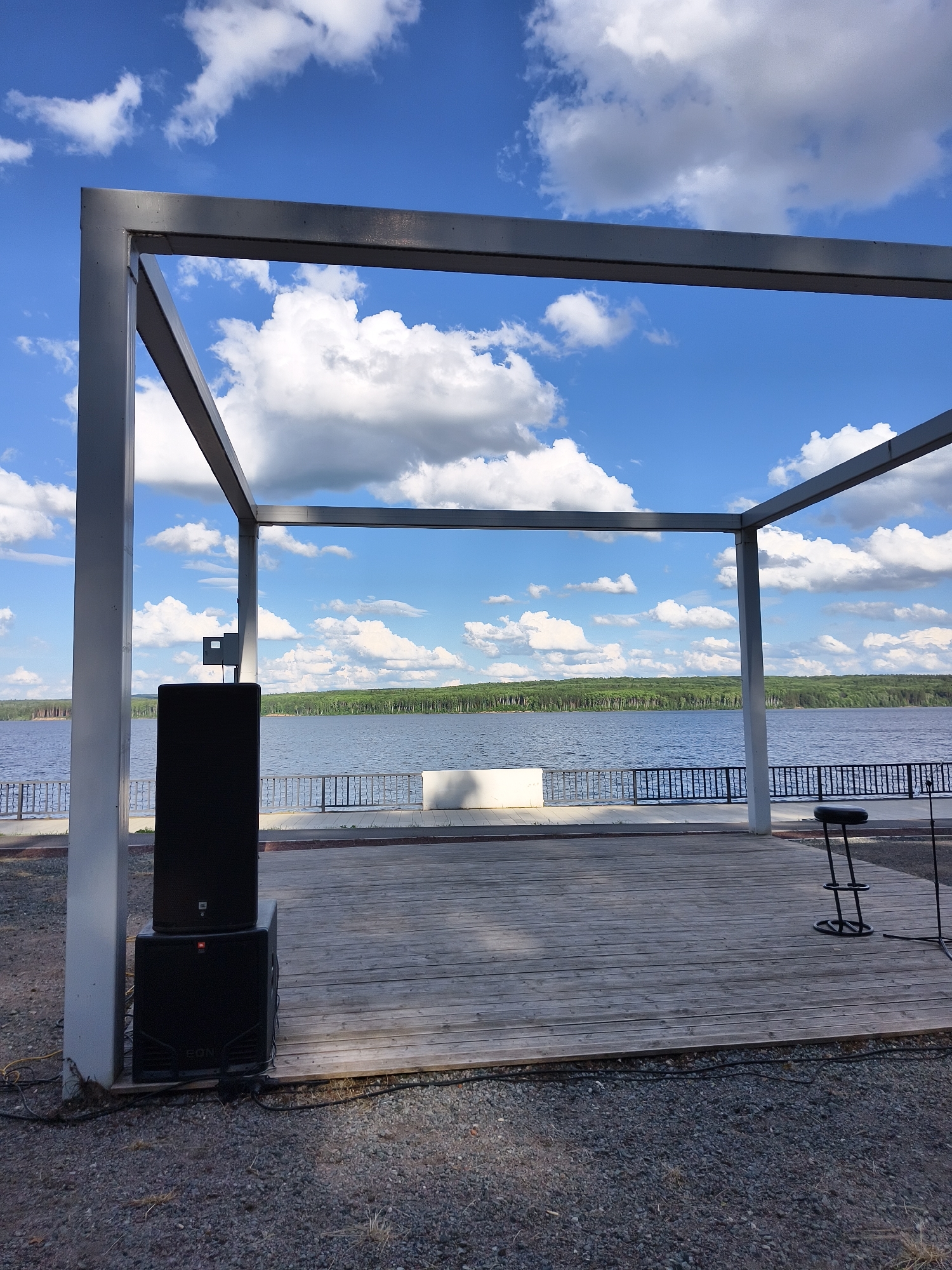 This is a landscape photograph taken outdoors on a clear day with bright sunlight. The scene depicts an outdoor stage area with a large, dark speaker system to the left. The stage is made of weathered wooden planks and is framed by a simple, modern, gray metal structure, likely a canopy or pergola.
In the background, a wide body of water, possibly a lake or river, stretches across the frame. The water is calm and reflects the bright blue sky dotted with fluffy white clouds. Beyond the water, a dense, green forest lines the horizon. A black metal railing runs along the edge of the stage area, separating it from the waterfront.
To the right of the stage, a black bar stool with a metal frame stands prominently. The overall atmosphere is serene and peaceful, suggesting a location designed for outdoor performances or gatherings. The time of day appears to be late morning or early afternoon, given the strong sunlight and clear sky. The weather is pleasant and sunny.
There is some visible text on the speaker system. The top speaker has a small, rectangular logo with "JBL" written inside. The bottom speaker has a larger logo which is a red circular logo with "JBL" inside, and below that "SON." The specific location is not identifiable from the image.