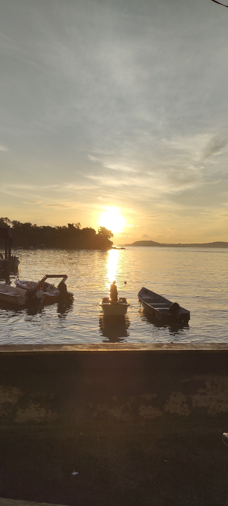 The image depicts a tranquil scene at the beach in Sabang, Indonesia, during sunset. The sun, a bright orb, is positioned low on the horizon, casting a warm, golden glow across the sky and the calm water. Wispy clouds streak across the sky, reflecting the sunlight.
In the foreground, a low concrete wall borders a grassy area. The main focus of the image is the calm body of water, likely the sea, with several boats visible. Three small boats are prominent in the middle ground. One white boat in the center contains a lone figure, silhouetted against the setting sun. Another boat, a darker, longer vessel, is positioned to the right of the central boat. To the left, a cluster of boats, including a white one with an orange frame and another smaller white boat, are moored.
In the background, a lush green island with dense trees is silhouetted against the bright sky. Further in the distance, rolling hills form the horizon. The water's surface is calm, with gentle ripples reflecting the colors of the sunset, creating a serene and picturesque atmosphere.