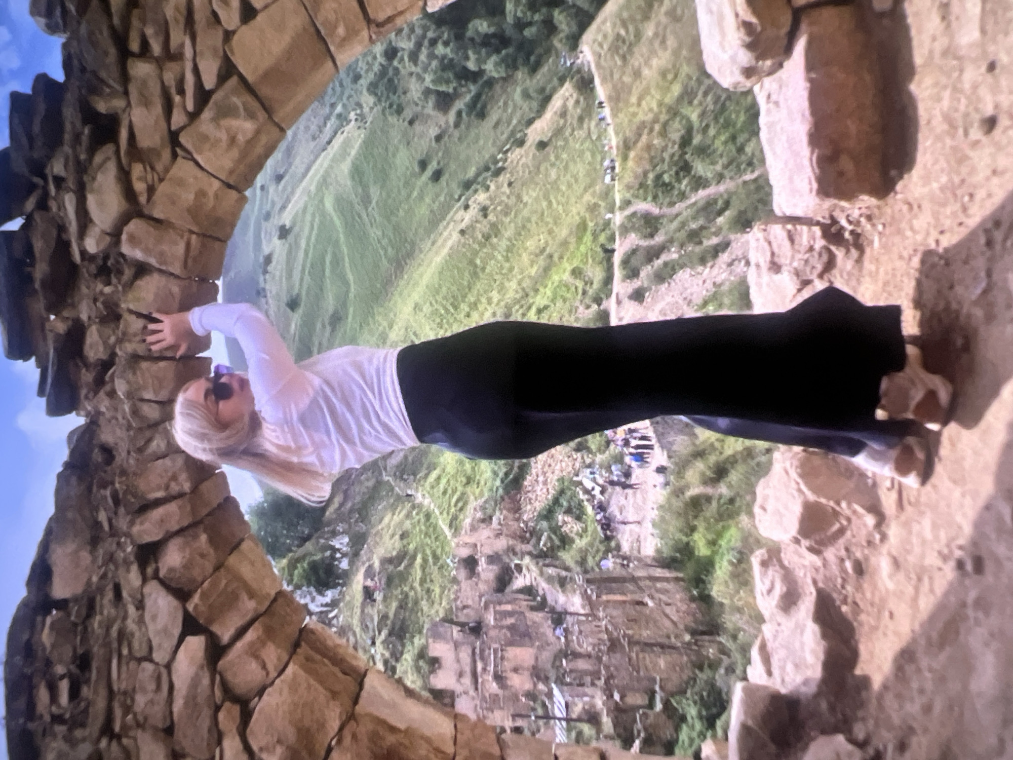 A full-body shot captures a blonde woman posing within a rustic stone archway, gazing out over a vast, green valley. The woman, positioned centrally, is dressed in a white long-sleeved top and dark, form-fitting pants or a long skirt. Her long, light-colored hair flows down her back, and sunglasses rest on top of her head. She holds onto the rough, irregularly shaped stones of the arch with both hands, her arms slightly extended, presenting a dynamic pose.
The archway itself is constructed from ancient-looking, weathered brown and grey stones, forming a sturdy, rounded frame. Beyond the arch, a dramatic landscape unfolds, characterized by steep, undulating green hills and deep ravines. Patches of darker vegetation, possibly small trees or bushes, are scattered across the slopes. Faint, winding dirt paths can be seen traversing the hills, and in the far distance, indistinct small structures or groups of people are visible, hinting at human activity within the expansive terrain.
The scene is brightly lit by natural daylight, suggesting clear skies and good weather. The ground around the archway is dry, rocky, and uneven, consistent with an outdoor historical or natural site. The image evokes a sense of exploration and appreciation for scenic vistas and ancient architecture. There is no visible text in the image.