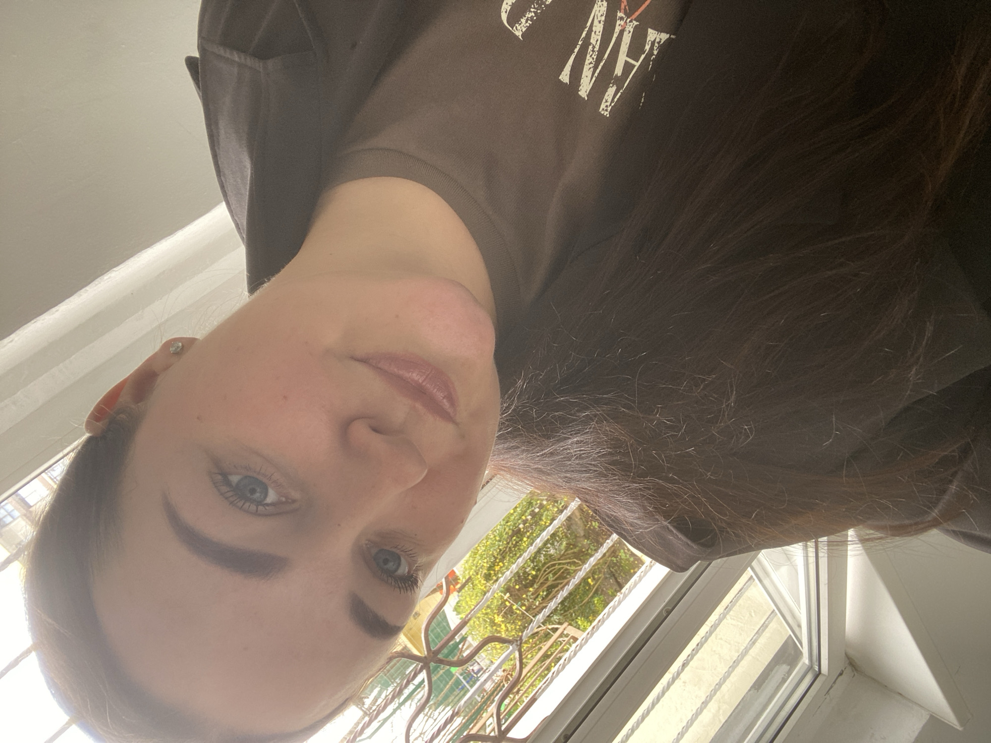 A low-angle, selfie-style portrait of a woman with fair skin and blue eyes. She is wearing a dark brown t-shirt with white lettering. Her dark brown hair is visible on the right side of the frame. The background shows a window with a metal railing, and a glimpse of green foliage outside. Based on the context, the location is possibly in Sochi, Russia.