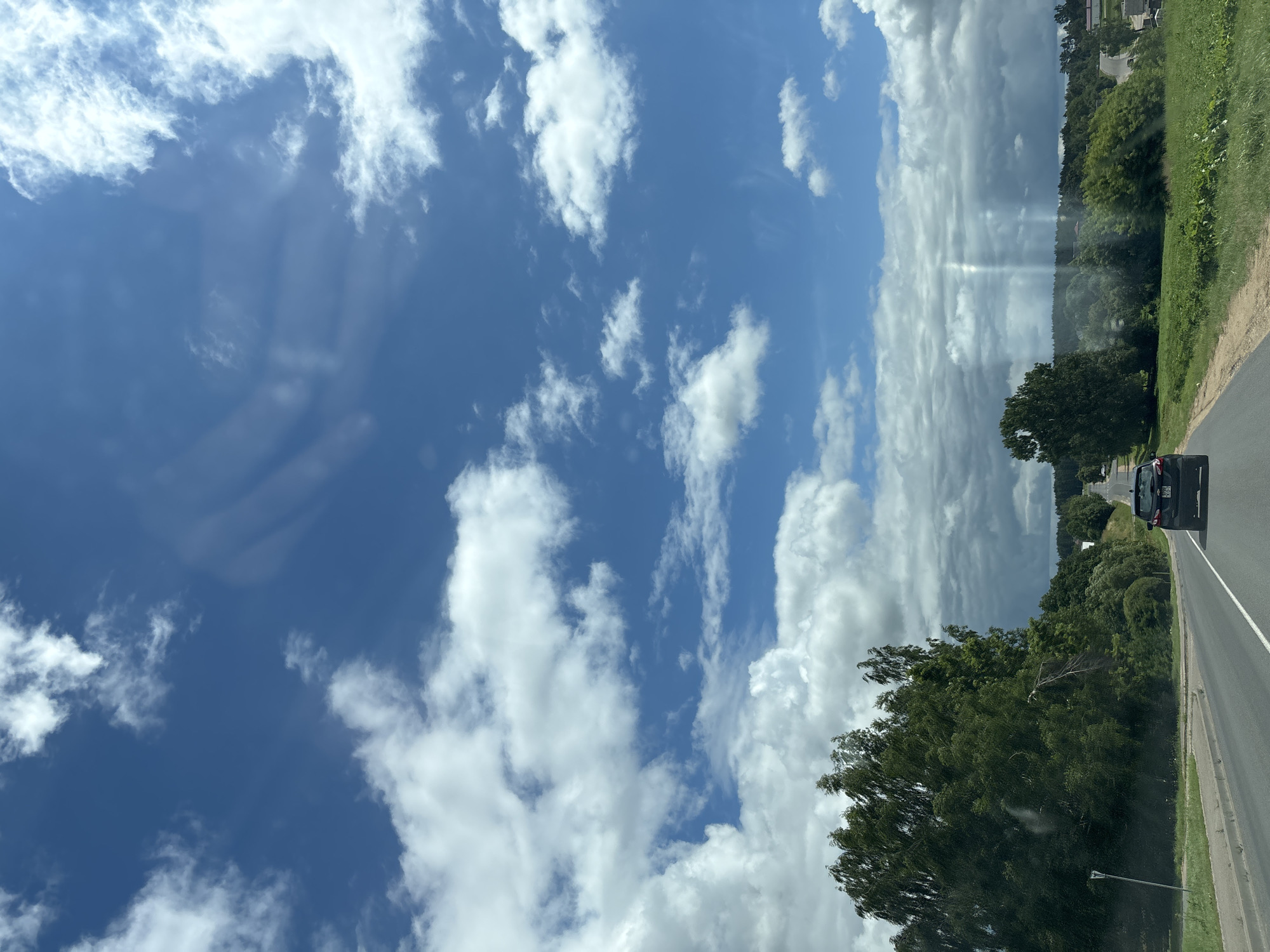 The image captures a view from a vehicle traveling along a road in the Kapylsky Selsoviet, Belarus. The sky is a vibrant blue with scattered white cumulus clouds, some of which are quite large and dense, obscuring portions of the sky. Sunlight beams are visible piercing through the clouds in the distance, creating a dramatic effect.
On the left side of the road, a dense line of green trees and foliage stretches along the roadside. A white signpost is partially visible among the trees. The road itself is paved with asphalt and has clear white lane markings.
A dark-colored car, possibly an SUV, is visible ahead on the right lane of the road, moving away from the viewer. The car's taillights are illuminated. The right side of the road is lined with more green vegetation, including bushes and grass. In the far distance, more trees and some scattered houses can be seen, indicating a rural or semi-rural setting within the Kapylsky Selsoviet. The overall impression is a sunny day with dynamic cloud formations, taken from a moving vehicle.
