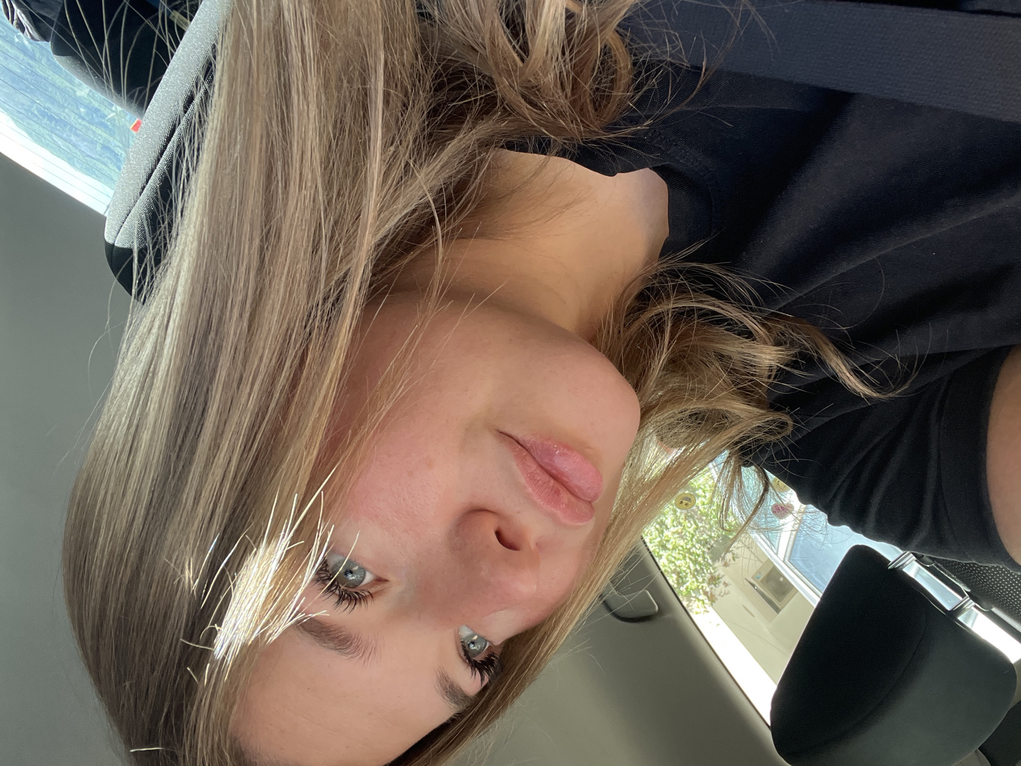 A young woman with blonde hair and blue eyes is in a car. She is wearing a black shirt. The interior of the car is visible. Outside the window, there are glimpses of greenery and buildings. The location appears to be Budva, Montenegro.