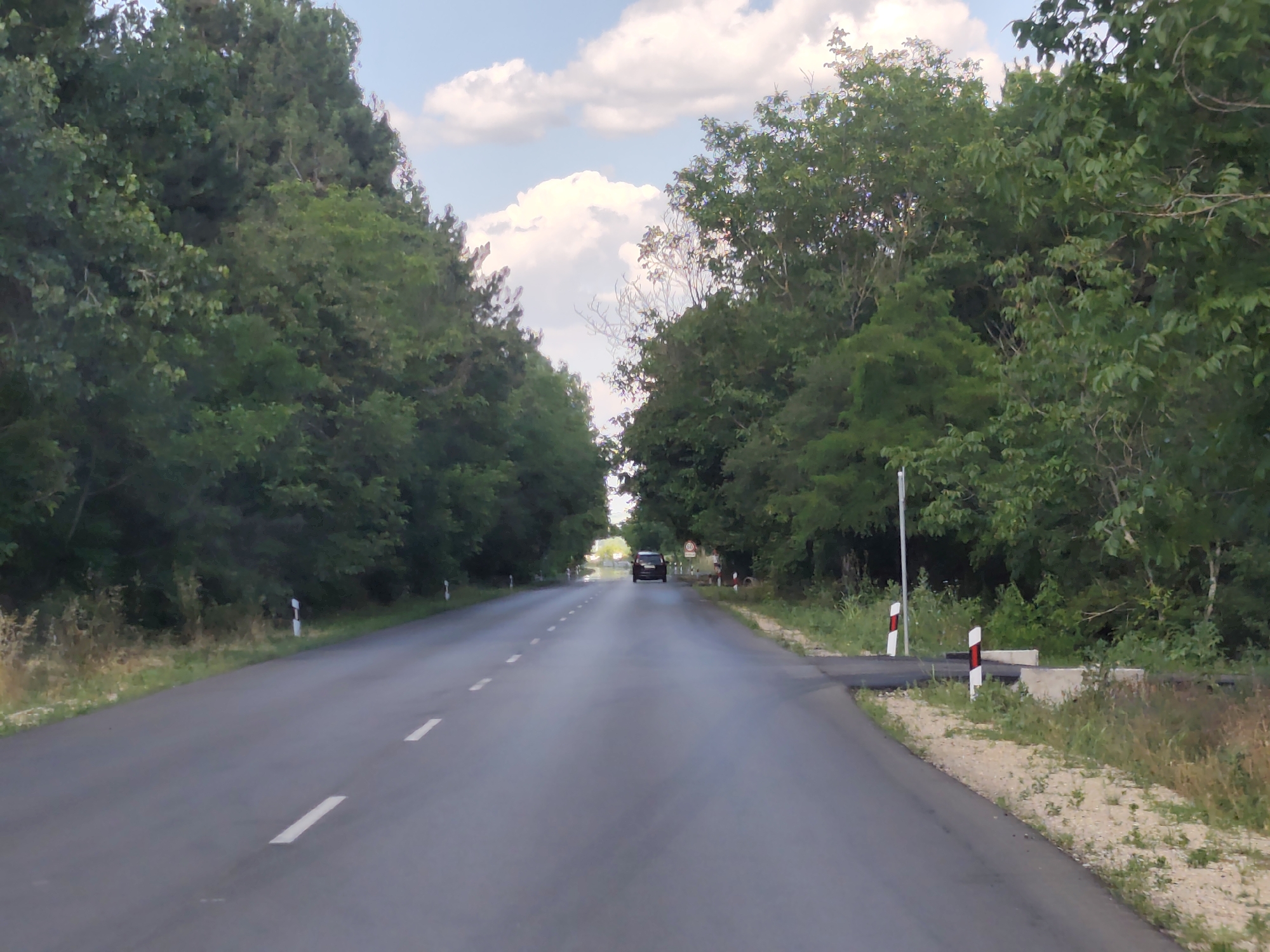 A dark-colored car drives down a paved road lined with trees on either side. The road has white dashed lines marking the lanes. The trees create a canopy over the road, with sunlight filtering through the leaves and onto the pavement. The sky above is blue with white, puffy clouds.
On the right side of the road, there are several white and red striped road markers, and some concrete barriers. A circular sign with red and white markings is visible on a pole in the distance. The scene appears to be set in a rural or natural environment, possibly a country road. The lighting suggests it is daytime, likely late afternoon given the angle of the sun and the warm tones in the sky. The overall mood is peaceful and serene, with the car appearing to be on a leisurely drive. There is no visible text in the image besides the road sign.