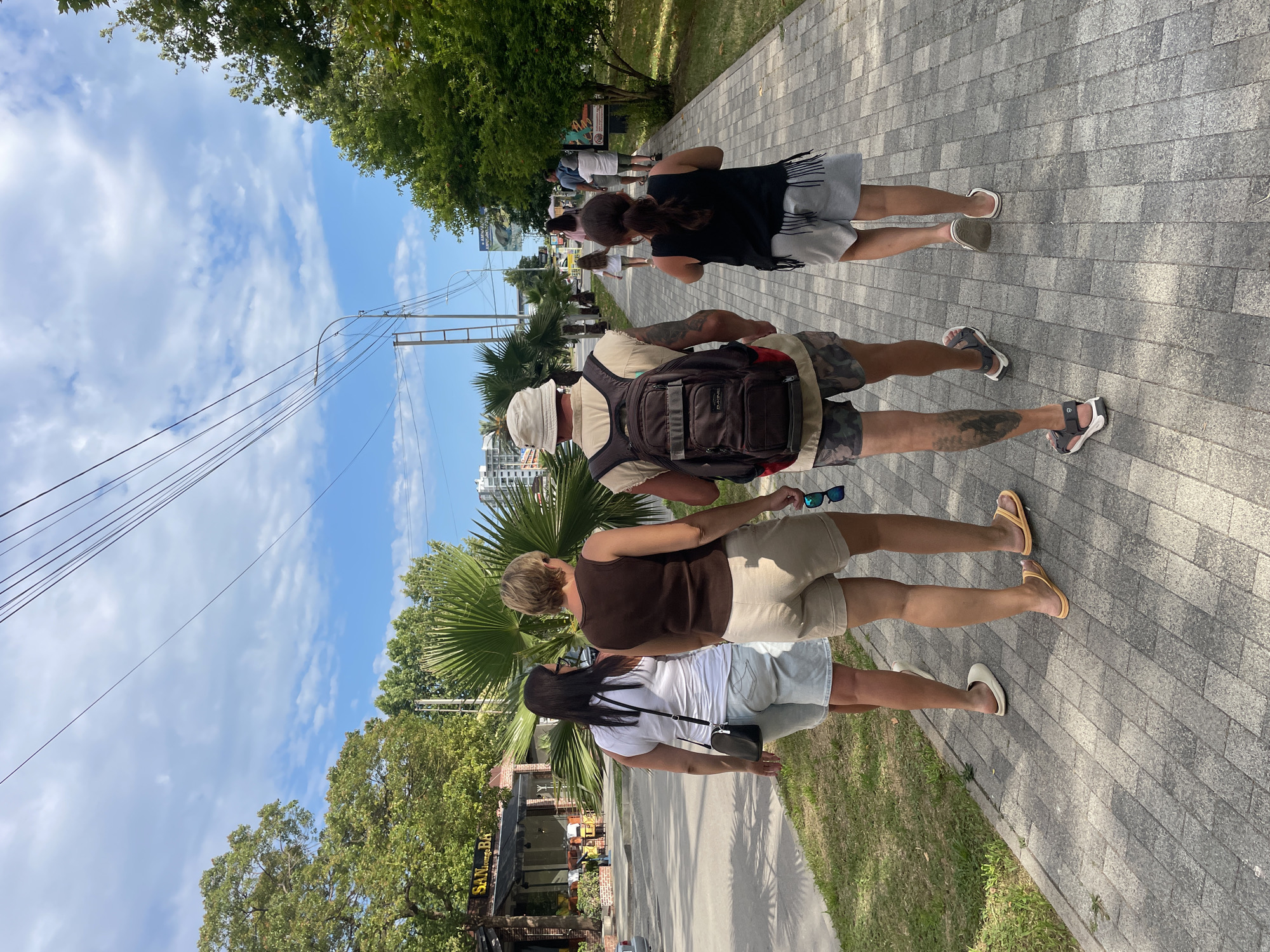 The image depicts a scene on a paved pedestrian walkway in Gagra, Abkhazia. Four individuals are walking away from the camera on the right side of the walkway. In the foreground, on the left side of the walkway, two women are walking. The woman closest to the camera is wearing a white tank top, light blue denim shorts, and white slip-on shoes. She has dark hair and is carrying a small black crossbody bag. The woman next to her is wearing a brown tank top, beige shorts, and yellow flip-flops. She has blonde hair and is holding a pair of blue sunglasses.
Further down the walkway, a man and a young girl are walking away from the camera. The man is wearing a light-colored hat, a tan vest over a light-colored shirt, camouflage shorts, and sandals. He has a dark backpack with red accents on his back and a tattoo on his right leg. The young girl is wearing a black tank top with fringe detail and grey shorts, and white flip-flops.
In the background, across the street from the walkway, is a commercial building with a sign that reads "SAN LOUNGE BAR". There are palm trees and other lush green trees lining the street and walkway. The sky is blue with scattered white clouds, and power lines are visible overhead. The overall environment suggests a warm, sunny day in a coastal or resort town.