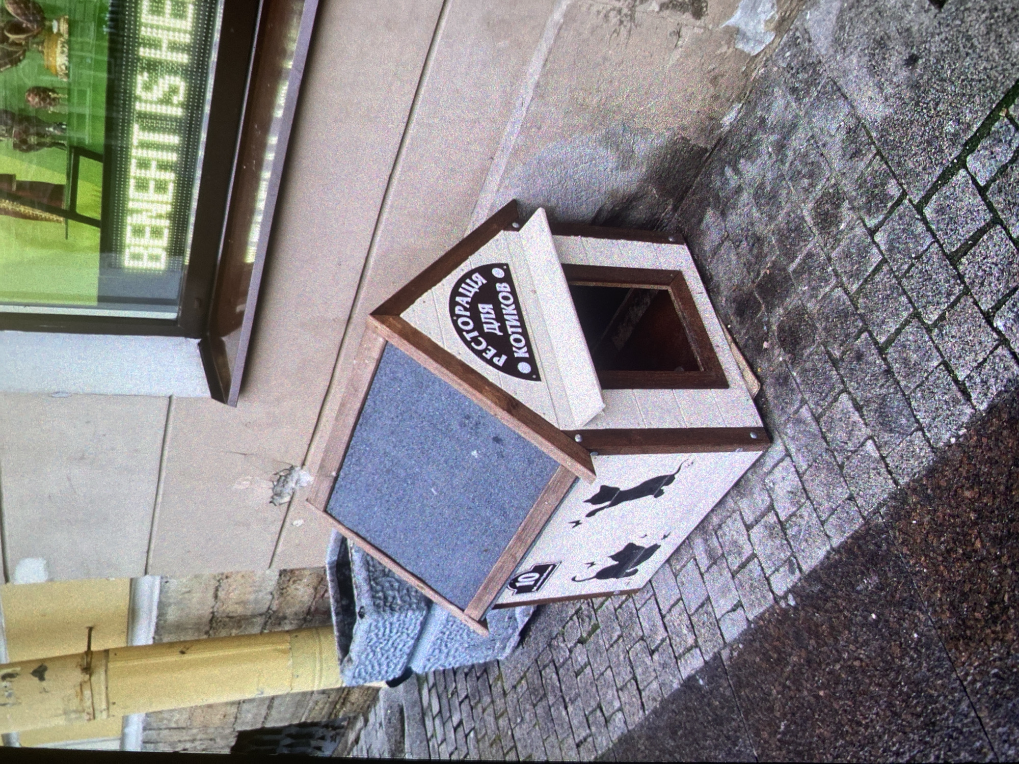 A cat house is seen on a brick sidewalk next to a building in Saint Petersburg, Russia. The cat house is white and brown with a gray floor and a sign in Russian that says "Restaurant for Cats." The cat house has a door that is open, and the floor is covered with a gray cloth. Two black cats are printed on the side of the house. The building next to the cat house has a window with a green sign that says "Benefit is He."