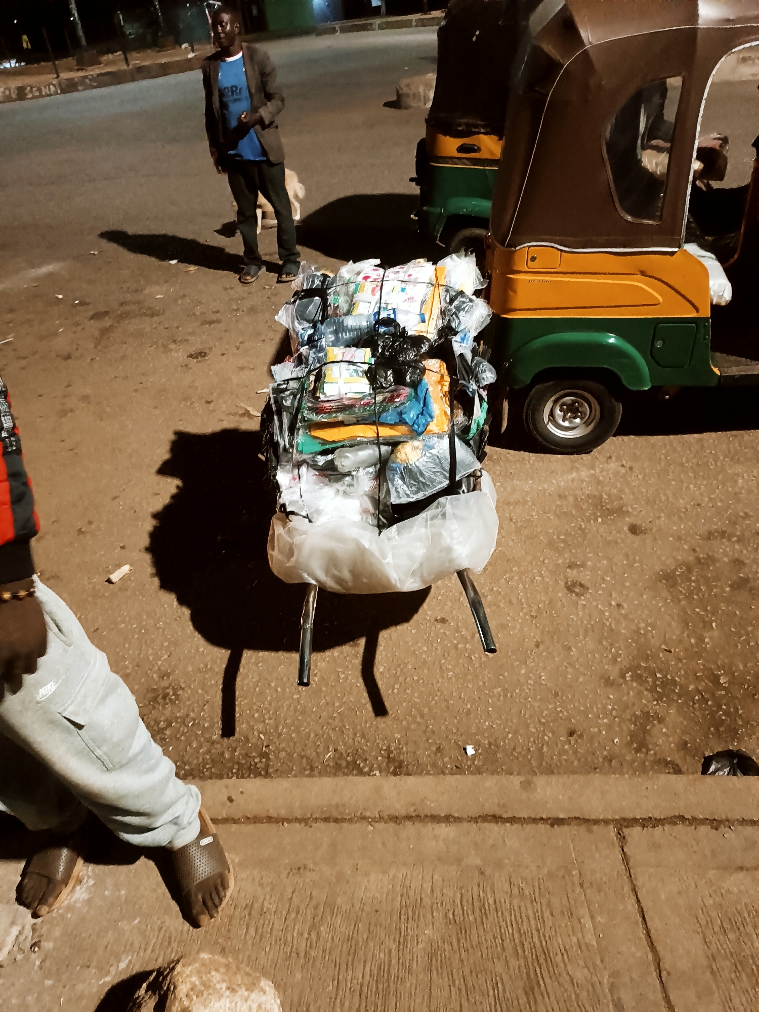 The image depicts a street scene in Bukuru, Nigeria. A man wearing a blue t-shirt under a brown jacket and dark pants stands on a paved surface. In the foreground, a densely packed load of various items, including textiles, plastic bags, and other unidentified objects, sits on a metal stand. To the right, a portion of a three-wheeled vehicle (likely an auto rickshaw or "keke") is visible, featuring a green and yellow color scheme. Another man, wearing grey sweatpants and sandals, partially obscures the left side of the image. The pavement shows signs of wear, with visible imperfections and debris. The time of day appears to be evening or night.