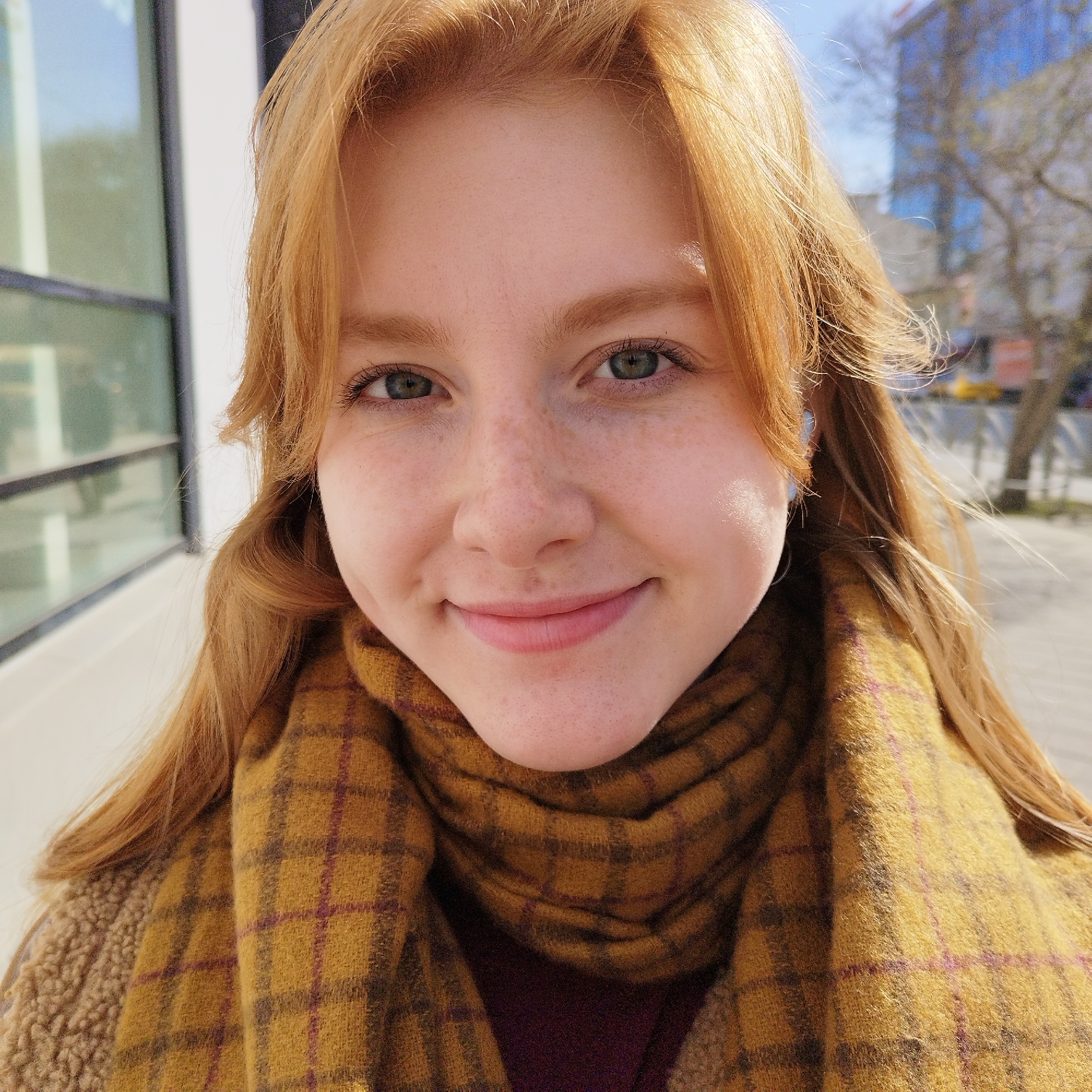 A close-up, eye-level portrait features a young woman with fair skin, freckles, and long, reddish-blonde hair. She has green eyes and is smiling gently at the camera. Her hair is parted in the middle and falls around her face and shoulders, with some strands on the right side appearing to be gently blown by a breeze.
She is wearing a thick, textured scarf in shades of mustard yellow with a prominent plaid pattern of darker yellow and burgundy lines. Underneath the scarf, hints of a brown, possibly fleece or teddy-textured, jacket or coat are visible. Small white wireless earbuds are discernible in her ears.
The setting appears to be outdoors during bright daylight, likely on a sunny day. The background is softly blurred but reveals architectural elements on the left, possibly a modern building with large windows reflecting the sky or surroundings. On the right, blurred trees and hints of other structures suggest an urban or suburban environment. The lighting creates highlights on her face and hair, particularly on the right side. No text is visible in the image.