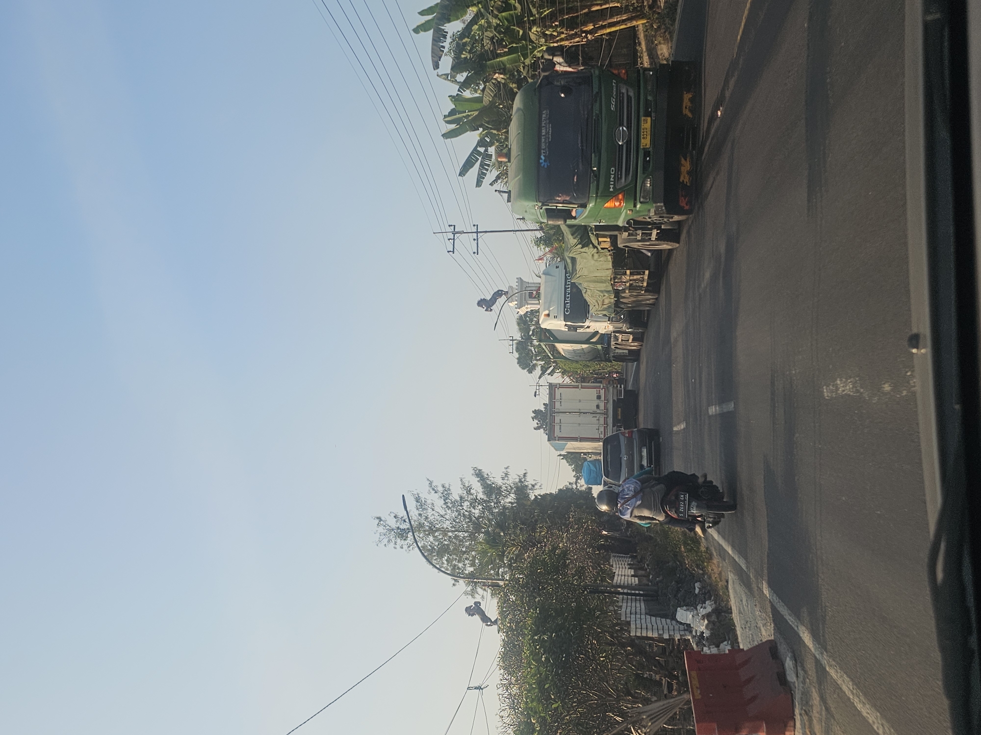 The image captures a traffic scene on a road in Semanding, Indonesia, likely during daylight hours given the clear sky. The perspective is from inside a vehicle, looking forward. A line of large trucks is traveling down the road, with a green Hino truck in the foreground on the right. The green truck has a license plate that reads "L 8319 UB" and has the text "PT DEWI SRI PUTRA" on its side. To the left of the green truck, other trucks are visible, including one with the text "CAKRAINT" and another white truck behind it. A motorcycle with two occupants is in the left lane, with the rider wearing a headscarf and the passenger also covered. On the left side of the road, there is vegetation, including trees and banana plants. A red traffic barrier is also visible on the left shoulder. In the distance, on the left, there is a white structure with a statue of a horse on top, connected to electrical wires. The road appears to be asphalt, with white lane markings.
