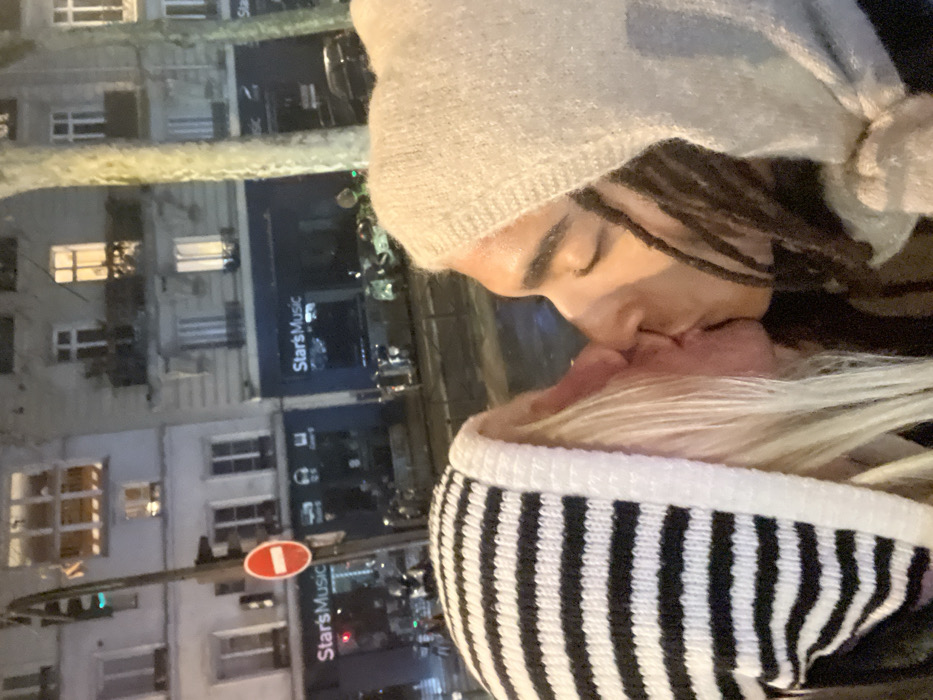 A couple kissing outdoors at night. The person on top wears a light gray beanie, dreadlocks visible underneath. The person below wears a white and black striped hoodie. A building with windows and a "StarsMusic" sign is visible in the background. A street sign with a red circle and horizontal white line is on the left. Street lights and other buildings contribute to the nighttime urban setting.