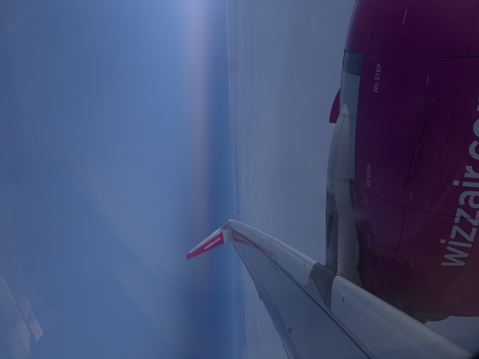 A photograph taken from the interior of an airplane shows the wing and engine of a Wizzair aircraft during flight. The wing, visible on the left side of the frame, is predominantly white and gray, with a pink section at the wingtip that bears the "wizzair.com" logo. The engine housing, located on the right side, is colored magenta, and also displays the word "wizzair.com" and the phrase "NO STEP." Outside the window, the sky appears predominantly blue, with a subtle gradient or possible cloud formation near the horizon. The location is not within the city of Uzundere, Turkey.