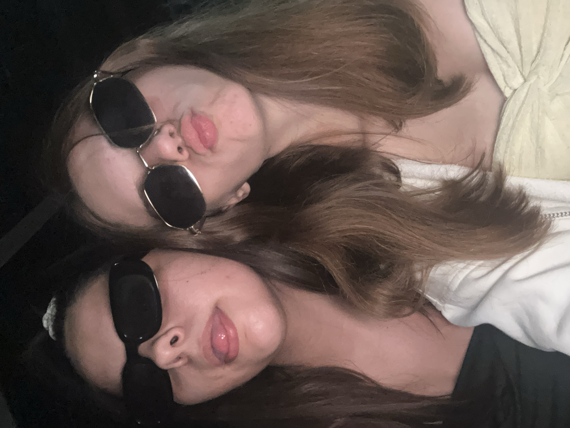 Two female subjects are positioned in a close-up, waist-up portrait taken in a low-light environment, likely indoors or at night, in the city of Unknown, Unknown. The background is uniformly dark and featureless. The lighting appears to be from a direct flash.
The subject on the left has long, dark brown hair. She wears opaque, black, rectangular sunglasses that cover her eyes. Her tongue is extended from her mouth. A light-colored, possibly textured hair tie is visible on the top of her head. She is wearing a dark, possibly off-the-shoulder or strapless garment.
The subject on the right has long, light brown hair. She wears opaque, gold-framed, octagonal sunglasses. Her lips are pursed into a pout. She is wearing a light yellow or cream-colored strapless or off-the-shoulder top with visible fabric texture. A white garment with a zipper is visible on her left side, partially covering her torso.
Both subjects are positioned closely together and appear to be looking towards the camera, consistent with a selfie.