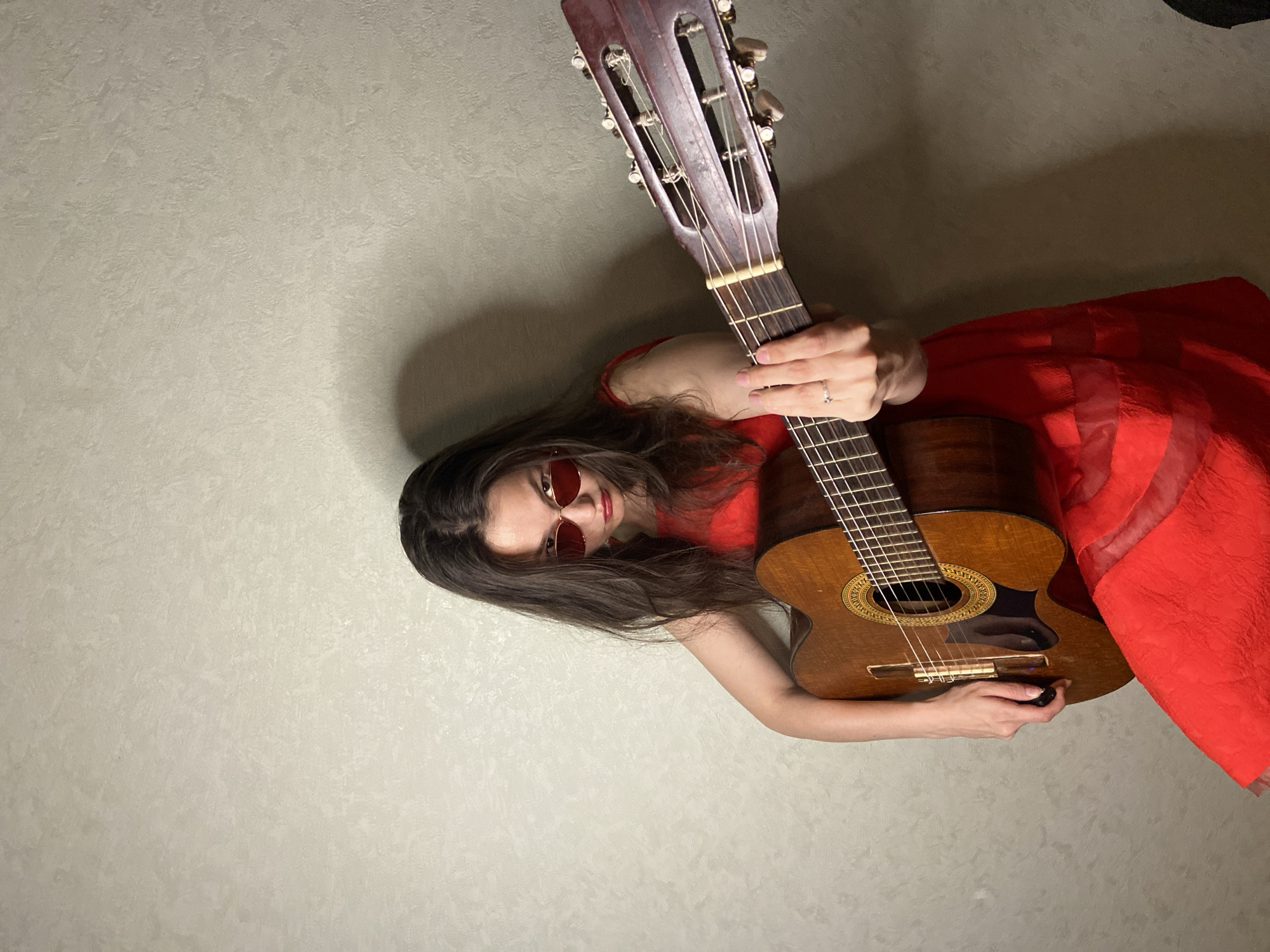 A young woman with long, dark hair and red-tinted sunglasses, wearing a bright red dress, plays a guitar. She is positioned in the center of the frame, leaning forward with a determined expression. Her left hand is on the fretboard, and her right arm is extended, suggesting she is either strumming or posing with the instrument. The guitar appears to be a classical or acoustic model with a wooden body. The background is a textured, light-colored wall with a shadow cast to the left of the woman. The lighting is dim, creating a moody atmosphere. There is no discernible indication of the city of Chaikovsky, Russia, within the image itself.