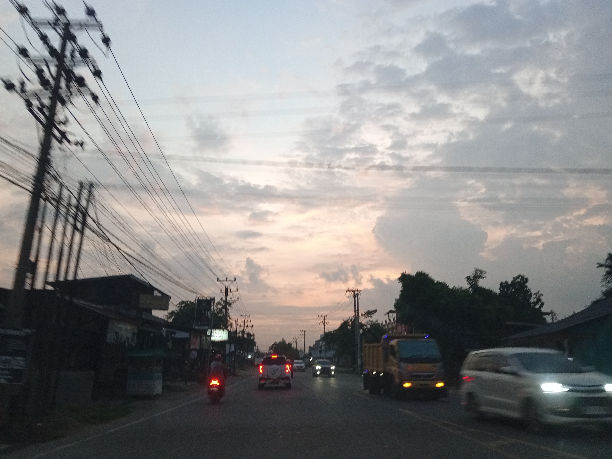 The image depicts a roadside scene at dusk in Payameuneng, Indonesia. A paved road runs through the frame, with a dark, overcast sky above. Utility poles with numerous wires are prominently positioned on the left and in the background, suggesting an urban or semi-urban environment.
On the left side of the road, there are several low-rise buildings, some appearing to be shops or small businesses, with awnings and signs visible. A single motorcycle, with its taillight illuminated, is traveling away from the camera.
In the center of the road, several vehicles are moving. A white SUV with its brake lights on is in front, followed by another car. Further down the road, a yellow truck is traveling in the same direction. On the right side of the road, a white car is approaching the camera, its headlights illuminated.
The overall impression is that of a typical evening commute or travel scene in a developing area, with active traffic and infrastructure present. The lighting suggests it is either sunrise or sunset.