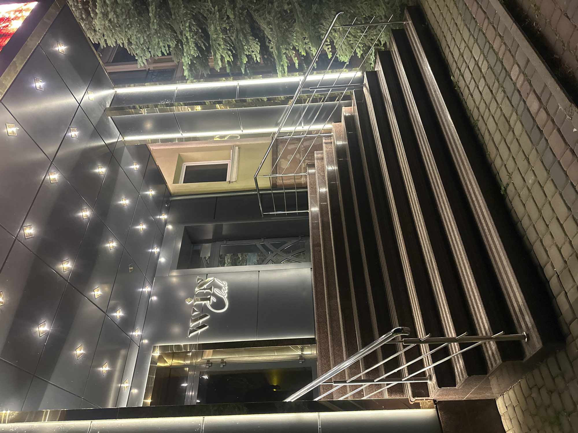 This is a nighttime photograph depicting the illuminated entrance of an establishment identified as "WIN Club" in Ternopil, Ukraine. A wide, multi-tiered staircase constructed of dark, polished stone with lighter horizontal accents leads upward to the main entrance. Stainless steel handrails with multiple horizontal bars flank both sides of the staircase.
The entrance features a prominent, dark-paneled canopy supported by several tall, reflective columns, some of which incorporate vertical strip lighting. The underside of the canopy is fitted with numerous square, recessed lights that cast a bright, localized illumination. The building's facade around the entrance is dark, possibly paneled, with reflective, metallic accents, including a prominent golden-toned frame above one of the glass doors. The "WIN Club" signage, composed of stylized white lettering and a golden crown motif, is affixed to the dark facade above the entrance. Two glass doors are visible behind the stairs; one features a distinct geometric design.
To the left of the entrance, a section of the building displays a light-colored wall, possibly pale green, with a single window. To the right, a large, dense evergreen tree, likely a conifer, is illuminated by the building's lights. The ground level in front of the stairs is paved with interlocking cobblestone-style pavers in varying shades of gray, brown, and reddish-brown. No individuals are present in the image.