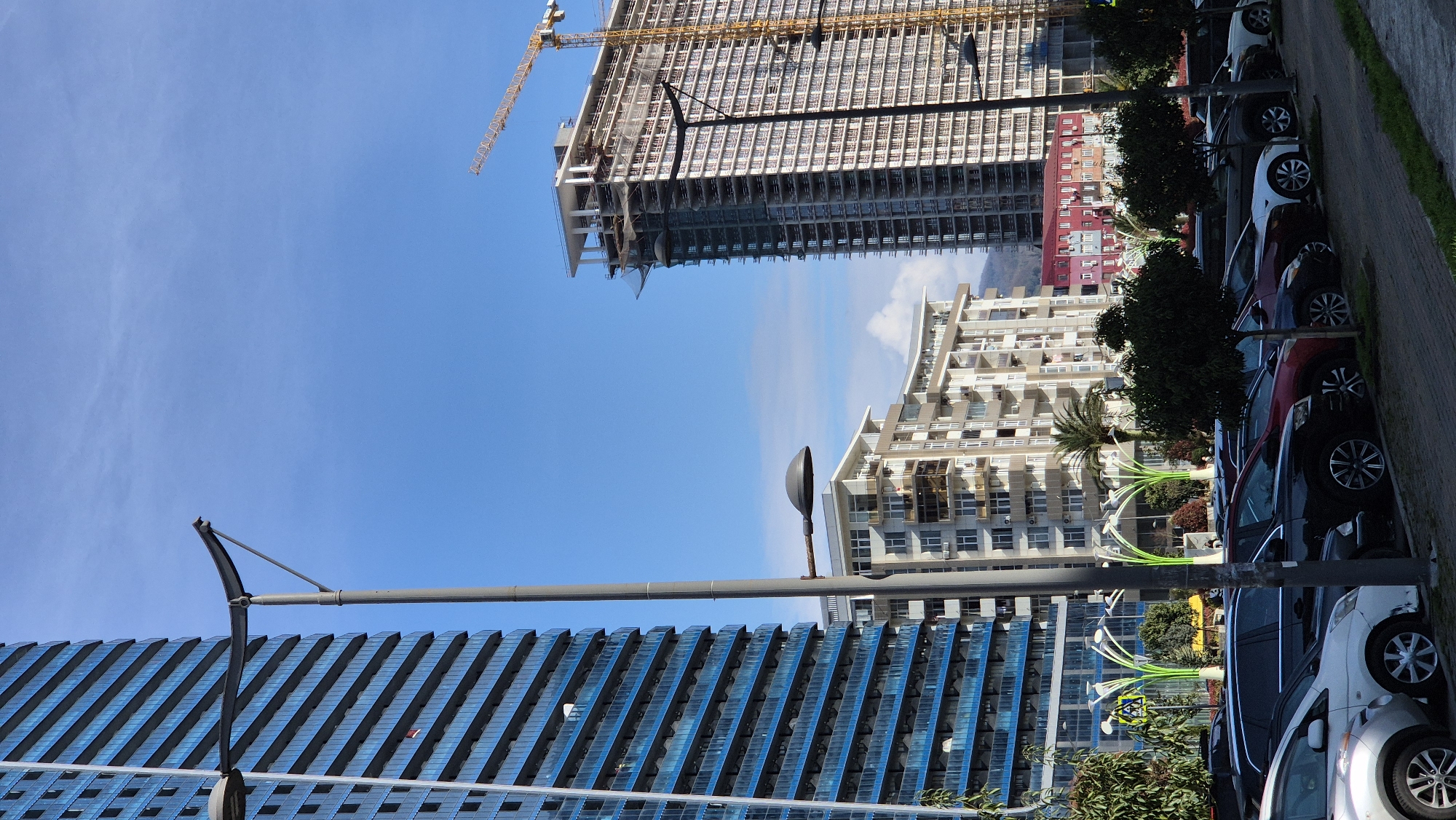 The image captures a scene in Batumi, Georgia, displaying a juxtaposition of architectural structures against a bright blue sky. A high-rise building under construction, marked by a yellow crane, stands tall, its skeleton exposed. Nearby, other multi-story buildings exhibit a more finished appearance. In the foreground, a line of parked cars runs parallel to the buildings, separated by lush greenery. Streetlights are also seen. The overall composition suggests an urban setting.
