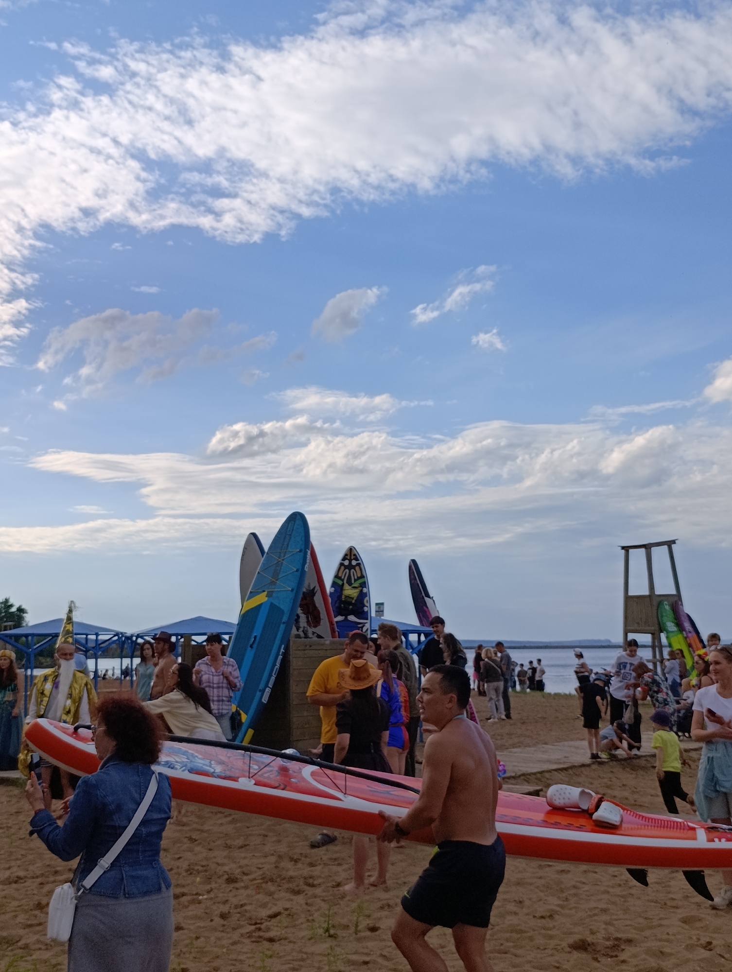 The image depicts a daytime scene on a sandy beach, likely in Chaikovsky, Russia, with a body of water and a cloudy blue sky in the background. A group of people are gathered, with several paddleboards visible, some standing upright and others being carried. In the foreground, a shirtless man is carrying a red and white paddleboard. To his left, a woman in a denim jacket and grey skirt is holding a phone, possibly taking a photo. Behind them, a man in a yellow shirt and hat is visible. To the left, a person dressed in a wizard-like costume with a yellow robe and pointed hat stands near a blue structure. Other individuals and paddleboards are scattered throughout the background, and a wooden lifeguard tower is situated on the right side of the frame. The environment suggests an outdoor event or gathering centered around water sports.