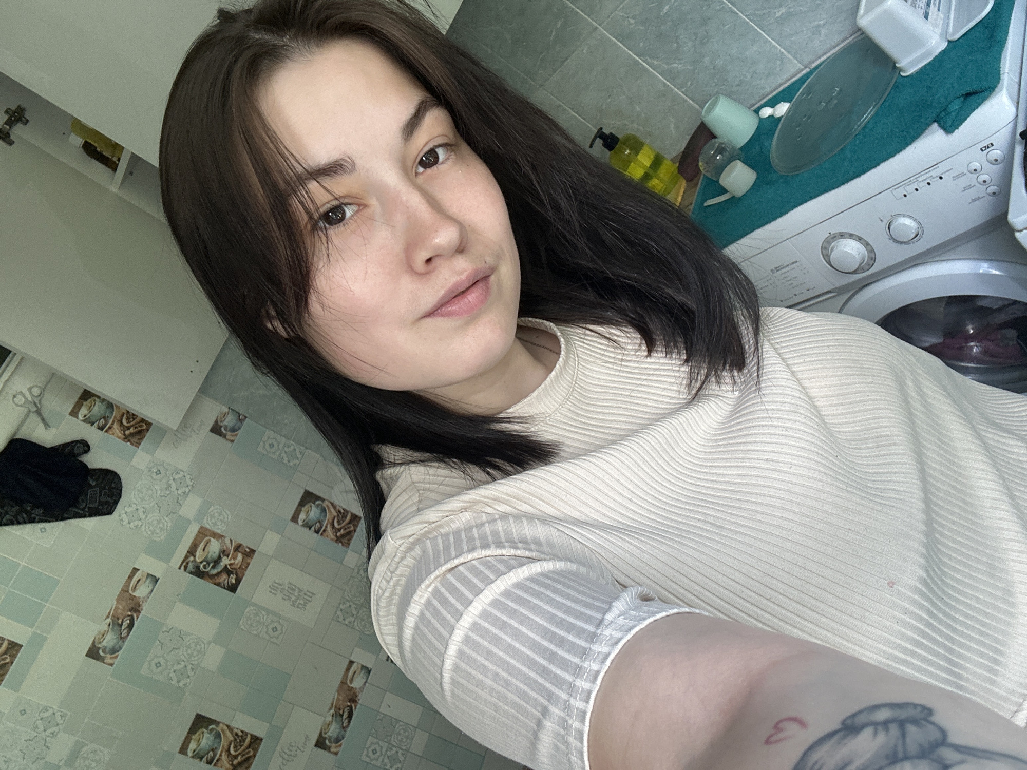 A young woman with dark hair takes a selfie in what appears to be a bathroom or laundry area. She is wearing a cream-colored, ribbed t-shirt. The background shows tiled walls with a coffee cup motif, a black oven mitt hanging, and some cleaning supplies. To the right, a washing machine is visible with a teal towel draped over it, holding several small containers and cups. There is a tattoo on the woman's left arm. The lighting is bright, likely from an overhead source. The location is not specified as Chaikovsky, Russia, but the provided context suggests it.