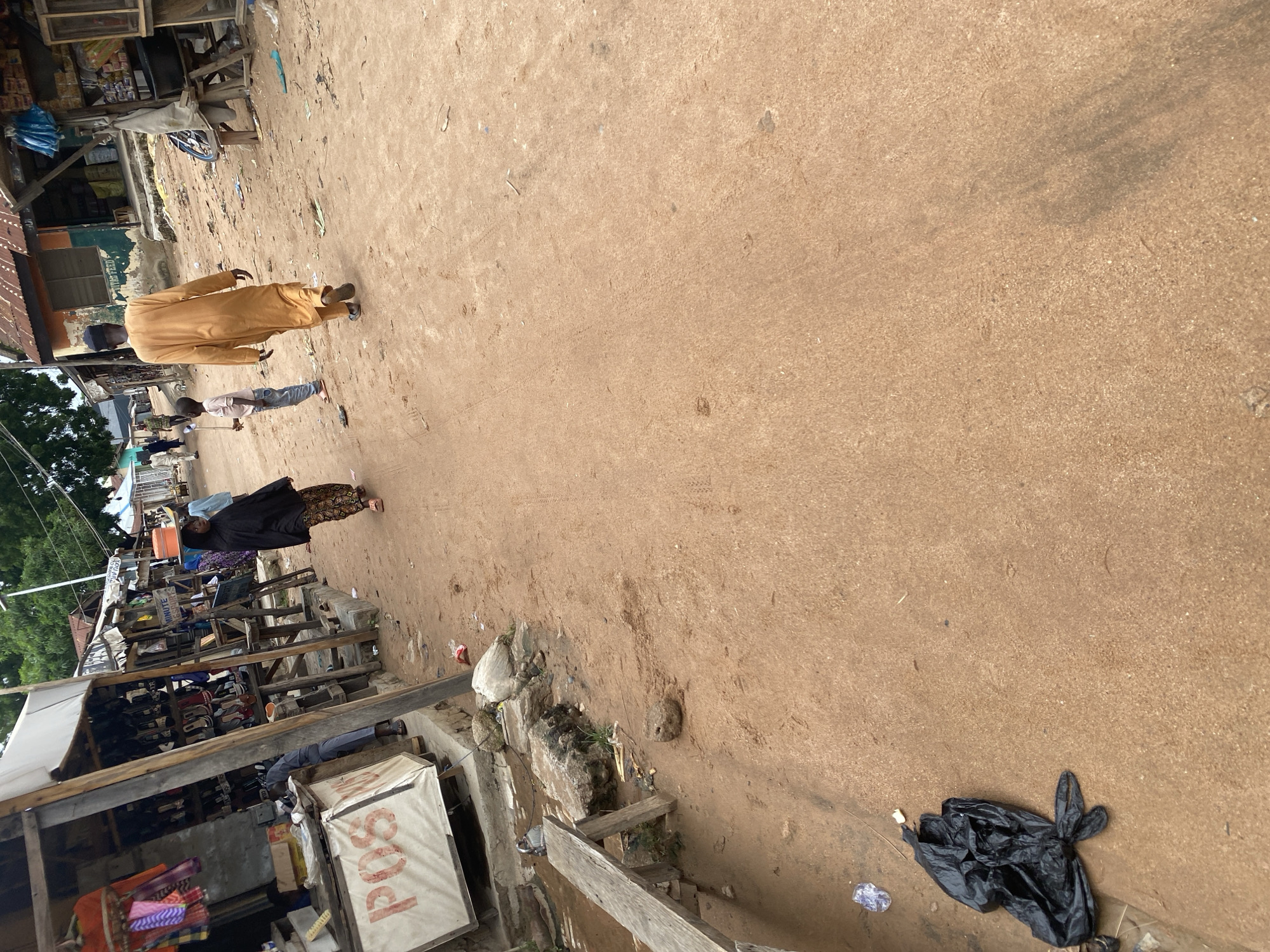 The media file depicts an outdoor scene in Jalingo, Nigeria. The setting appears to be a bustling marketplace with a dirt or unpaved road. Several individuals are visible. A person in a long, tan-colored garment walks away from the camera, while another person, possibly a child, in a lighter colored shirt and jeans, stands nearby holding a stick. Further down the road, another individual in a dark outfit with a patterned skirt is walking.
There are numerous market stalls and structures made of wood and corrugated metal lining the sides of the road. Some stalls display goods such as textiles, shoes, and packaged items. A white tarp covers part of a stall, and on it, the letters "POS" are visible in red. Scattered debris and a black plastic bag lie on the ground, suggesting a busy, possibly less-than-pristine environment.
The lighting suggests it is daytime, likely with sunny weather given the clear visibility and the lack of shadows indicating overcast conditions. The overall impression is a snapshot of daily life and commerce in a Nigerian town.