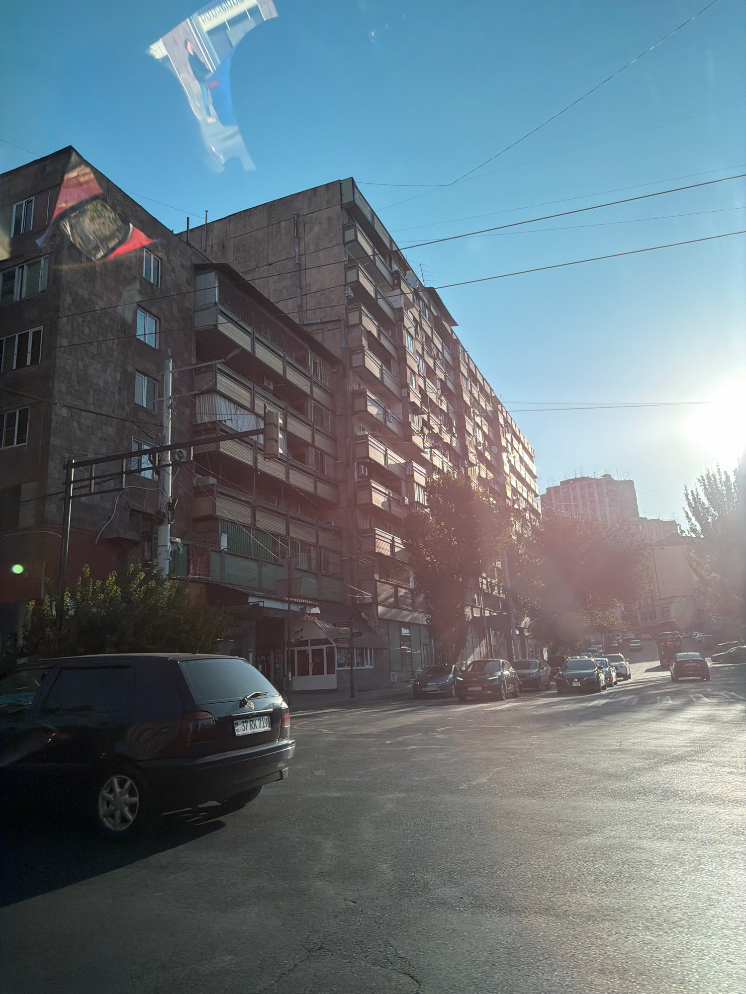 A black station wagon is parked on the right side of a street in Yerevan, Armenia, with its rear facing the viewer. The car's license plate reads "37 RK 719". In the background, a large, multi-story apartment building with balconies lines the street. Several other cars are visible on the road, and the sun is shining brightly, casting long shadows. The sky is clear and blue. The image appears to be taken from inside another vehicle, as evidenced by reflections on the windshield.