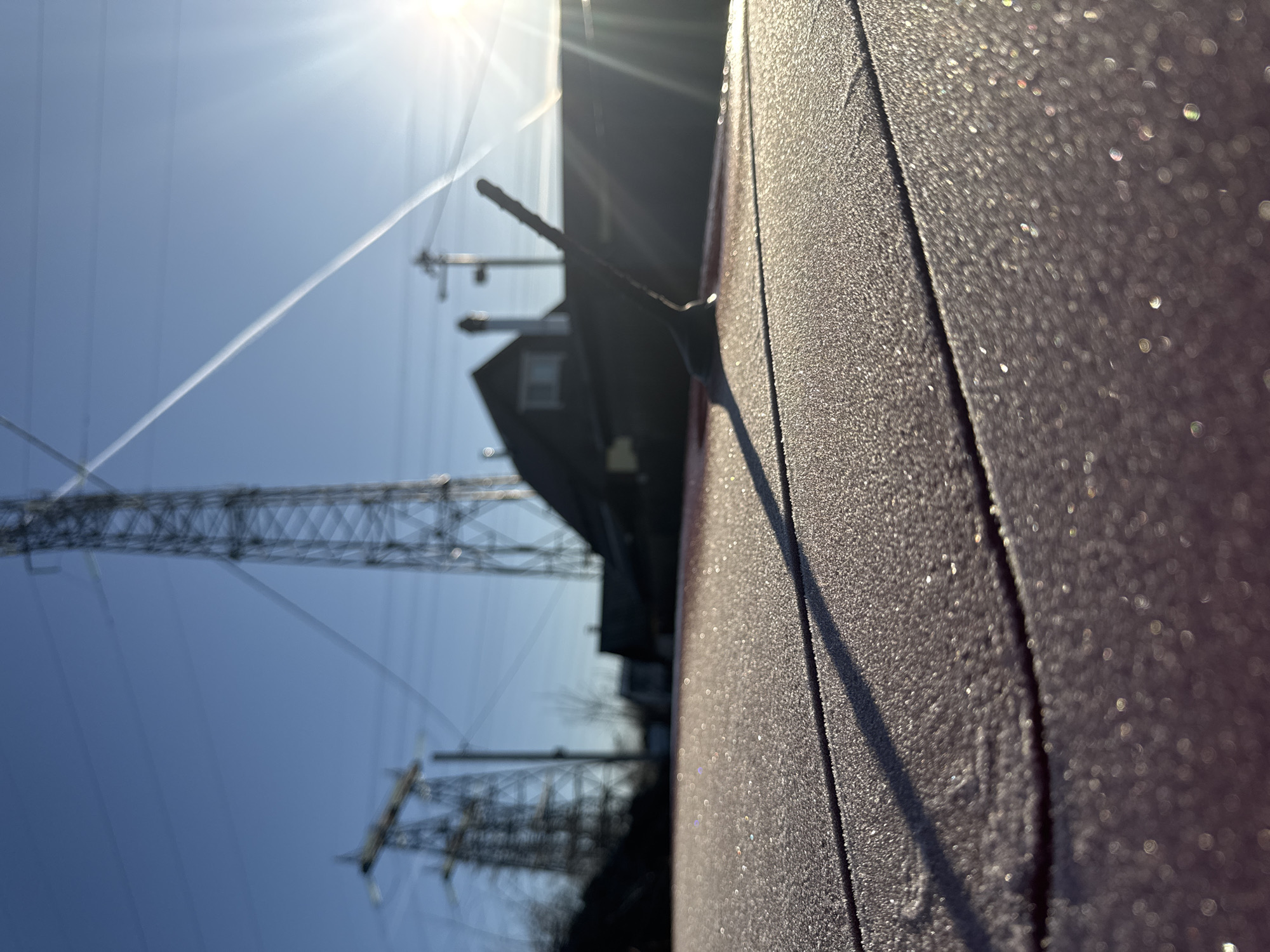 A low-angle, rotated view captures a frosty surface in the foreground, possibly a car's exterior. The frost is illuminated by the sun, creating a shimmering effect. In the background, a power transmission tower and lines are visible against a clear blue sky. A contrail cuts across the sky. The silhouette of a building or structure is positioned behind the tower. Based on the presence of power transmission towers and the prompt's specifications, the image suggests an urban or industrial area in or around Chaikovsky, Russia. The scene depicts a cold, sunny morning where frost has formed on surfaces.