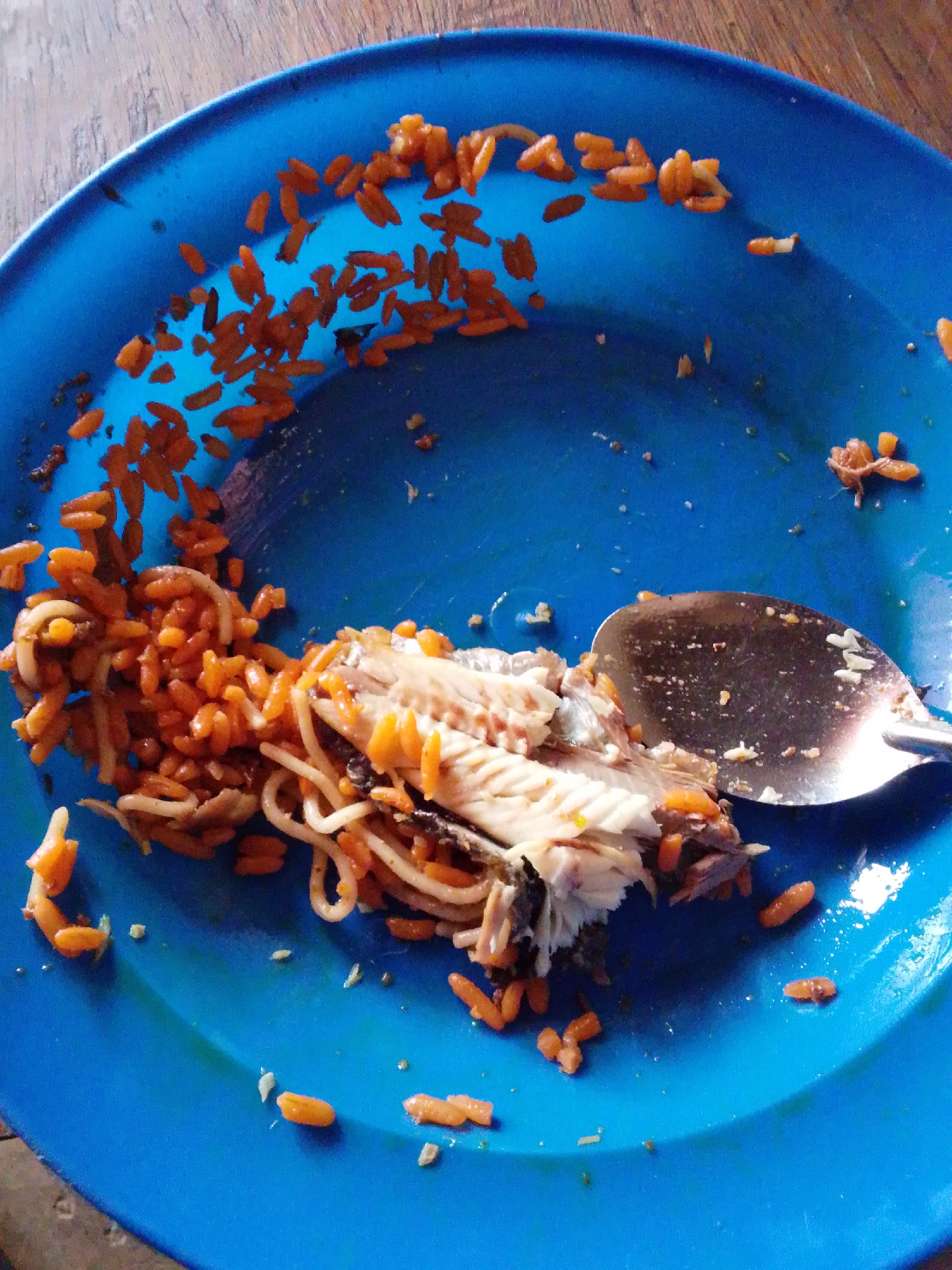 A close-up, high-angle shot shows a blue plate with a portion of a fish and rice dish. The plate is on a wooden surface. The dish appears to be mostly eaten, with a significant amount of rice scattered around the plate. A spoon is resting on the right side of the plate, partially covering the remaining food. The fish is deboned and appears to be white and flaky. The rice is small, oval-shaped, and orange in color, possibly seasoned with paprika or tomato sauce. Some strands of pasta are mixed in with the rice and fish. The overall impression is of a simple meal that has been consumed.