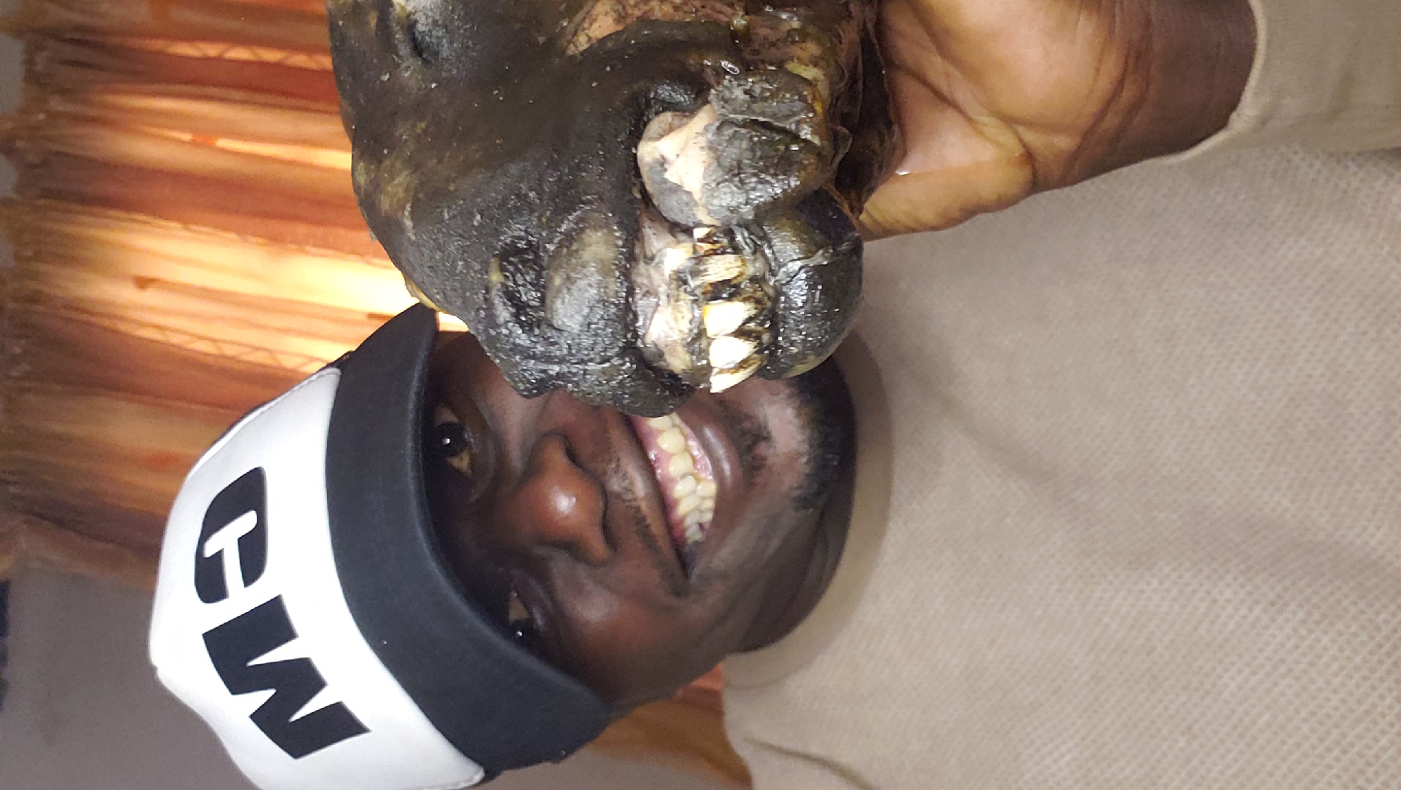 A man wearing a white and black baseball cap with "M3" printed on the front, and a tan shirt, smiles at the camera while holding a dark, wet, animal head with prominent teeth. The head appears to be that of a goat or similar livestock. The background consists of orange-brown curtains. The image was taken in Sabon Tasha, Nigeria.