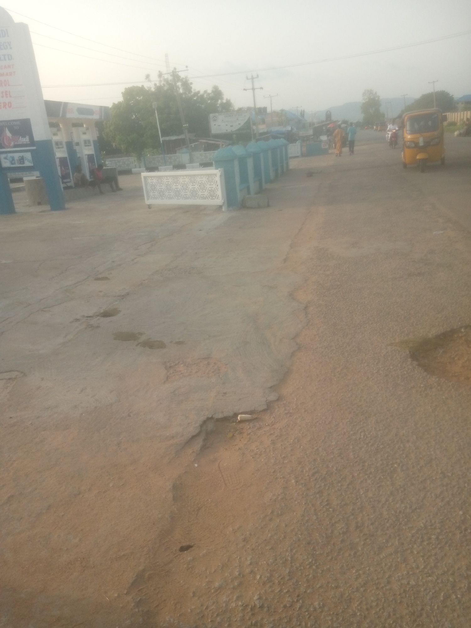 The image depicts an outdoor scene in the city of Jalingo, Nigeria. In the foreground, a rough, textured asphalt road occupies the majority of the frame, with areas of lighter colored sand and debris on the left. To the left of the road, a white gas station sign with blue accents is visible, displaying text such as "STRATEGY LTD." and "WELCOME TO Audi". Adjacent to the sign are gas pumps and a small building. Further down the road, a white decorative gate with a geometric pattern is present. In the midground and background, several people are walking, and a yellow tricycle-like vehicle is driving. Trees and buildings can be seen in the distance, along with utility poles and overhead wires. The sky is overcast and light-colored, suggesting it might be early morning or late afternoon.