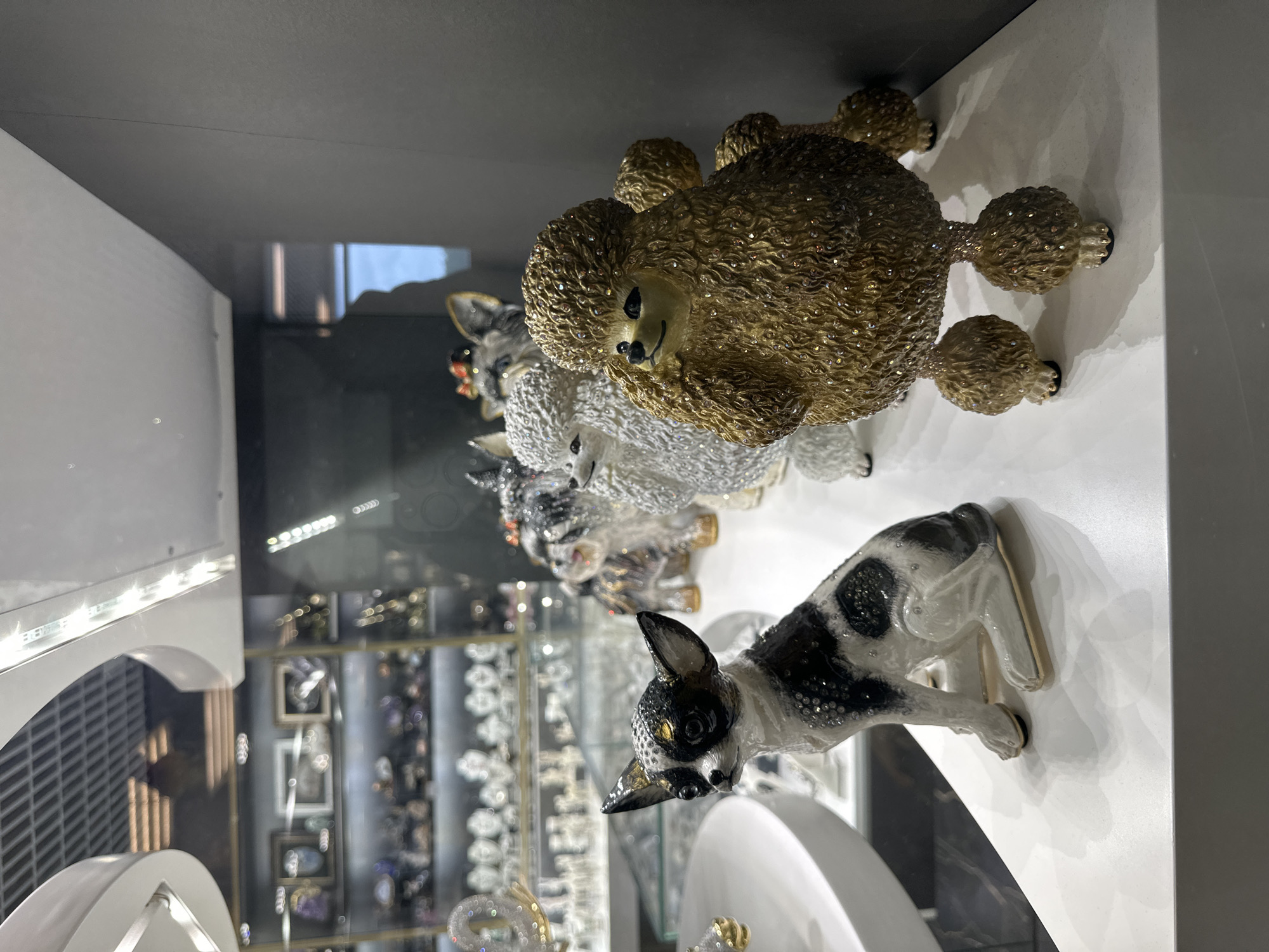 Inside a brightly lit display case, several animal figurines are visible. The foreground features a porcelain dog with black and white markings, positioned as if peering into the camera. Behind it are three poodle figurines, each rendered in a different color – gold, white, and silver. The figurines are densely covered in glittering decorations. The backdrop of the display case is a blurred array of objects, possibly jewelry, indicating this is a retail setting. The scene is located in the city of Saint Petersburg, Russia.