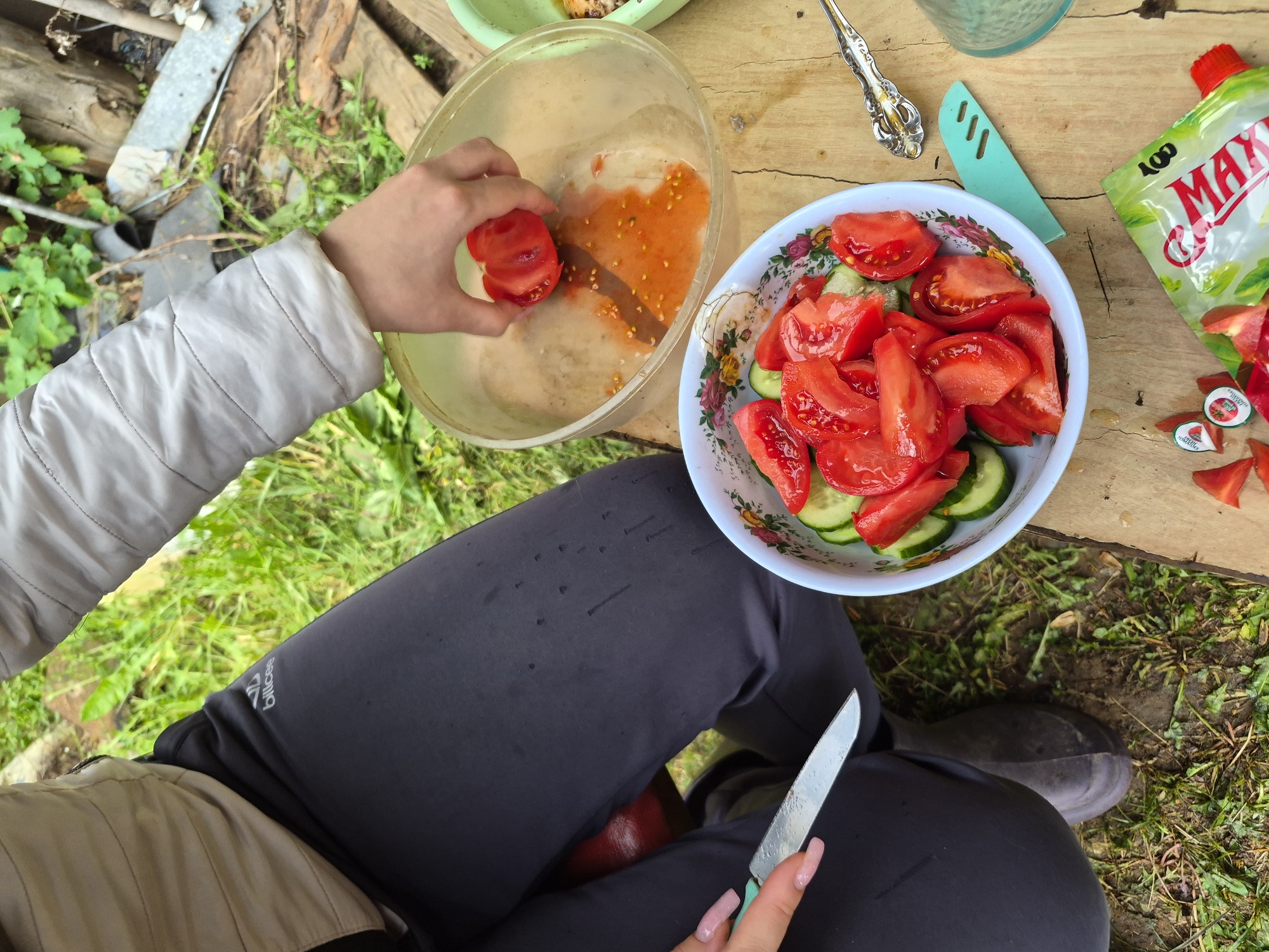 In Kharnavy, Russia, a person is engaged in food preparation outdoors. The scene is set on a worn wooden surface, with an unkempt grassy and dirt area, interspersed with various discarded wooden and metal fragments, serving as the background. The individual is wearing a light-colored, long-sleeved jacket and dark grey or black athletic-style pants, featuring a white "billcee" logo on the left thigh. A dark boot is visible on the left foot.
The person's right hand, displaying long, light-colored manicured fingernails, holds a large, cut red tomato over a clear plastic bowl. A significant amount of tomato juice and seeds has collected at the bottom of this bowl. The left hand holds a silver knife with a light blue-green handle, which has visible water droplets on its blade.
On the wooden surface, a white bowl adorned with a floral pattern (roses) is filled with freshly sliced red tomatoes and green cucumbers, arranged as a salad. Adjacent to this bowl are a decorative silver spoon, a light blue-green plastic serving utensil, and a partially visible clear drinking glass. A green and white flexible pouch, with "MAX" and "100" visible in its branding (suggesting mayonnaise or a similar condiment), lies open on the wood, surrounded by small pieces of cut tomato and two small, red-capped packets. The overall activity depicts the assembly of a fresh vegetable salad.